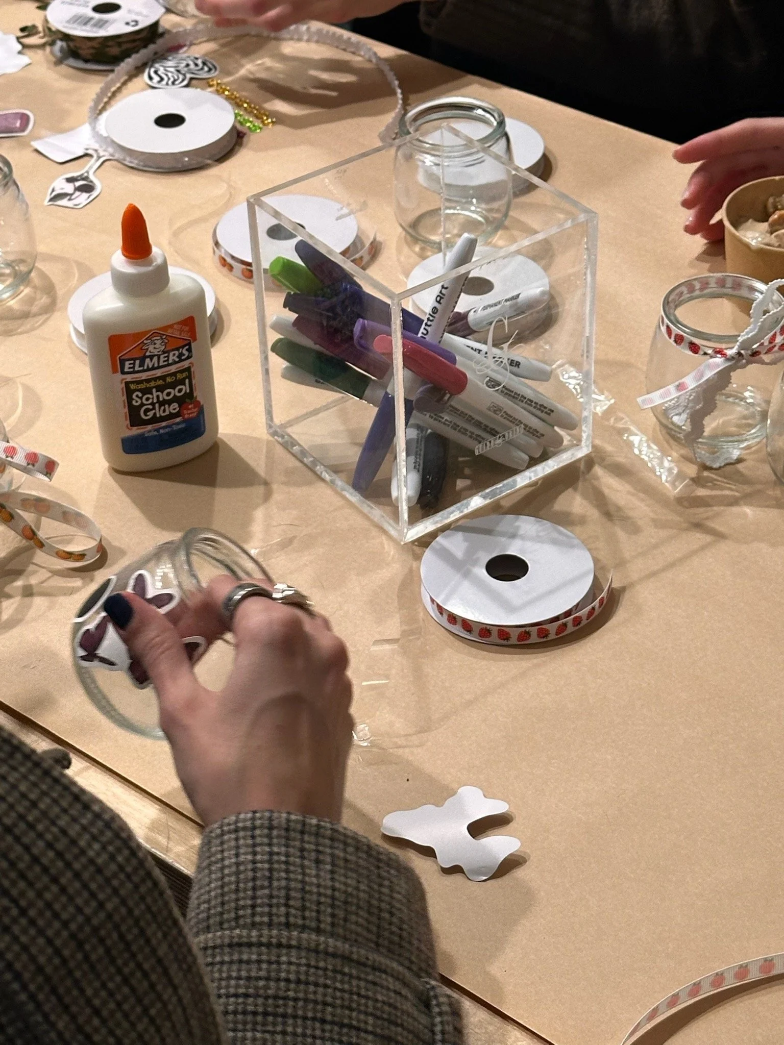 Crafting table with colorful markers, glue, ribbon spools, glass jars, and cutouts for a craft or decoration project, with hands working on the project.