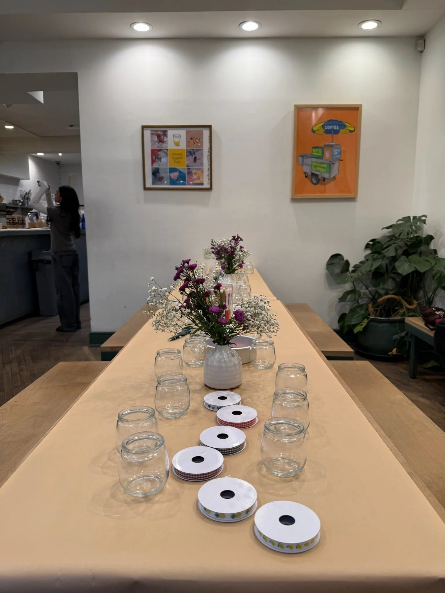 A long table set with a flower arrangement and empty glasses, surrounded by rolls of ribbon in a brightly lit room with white walls, framed artwork, and a large leafy plant.