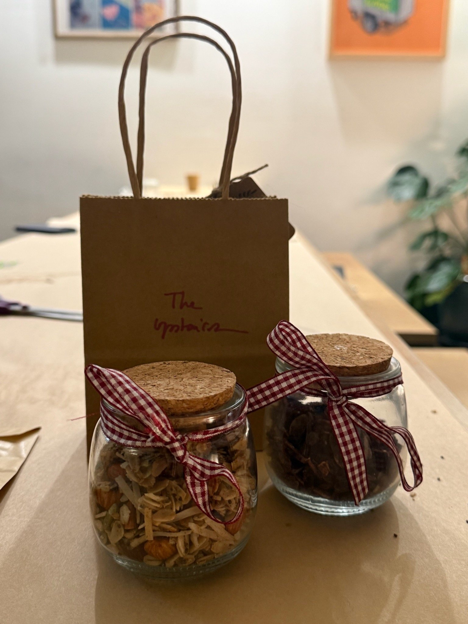 Two glass jars with cork lids and red checkered ribbons tied around the necks, filled with dried herbs or tea leaves, and a paper bag with a handwritten label that says 'The UpTopia' on a wooden table.
