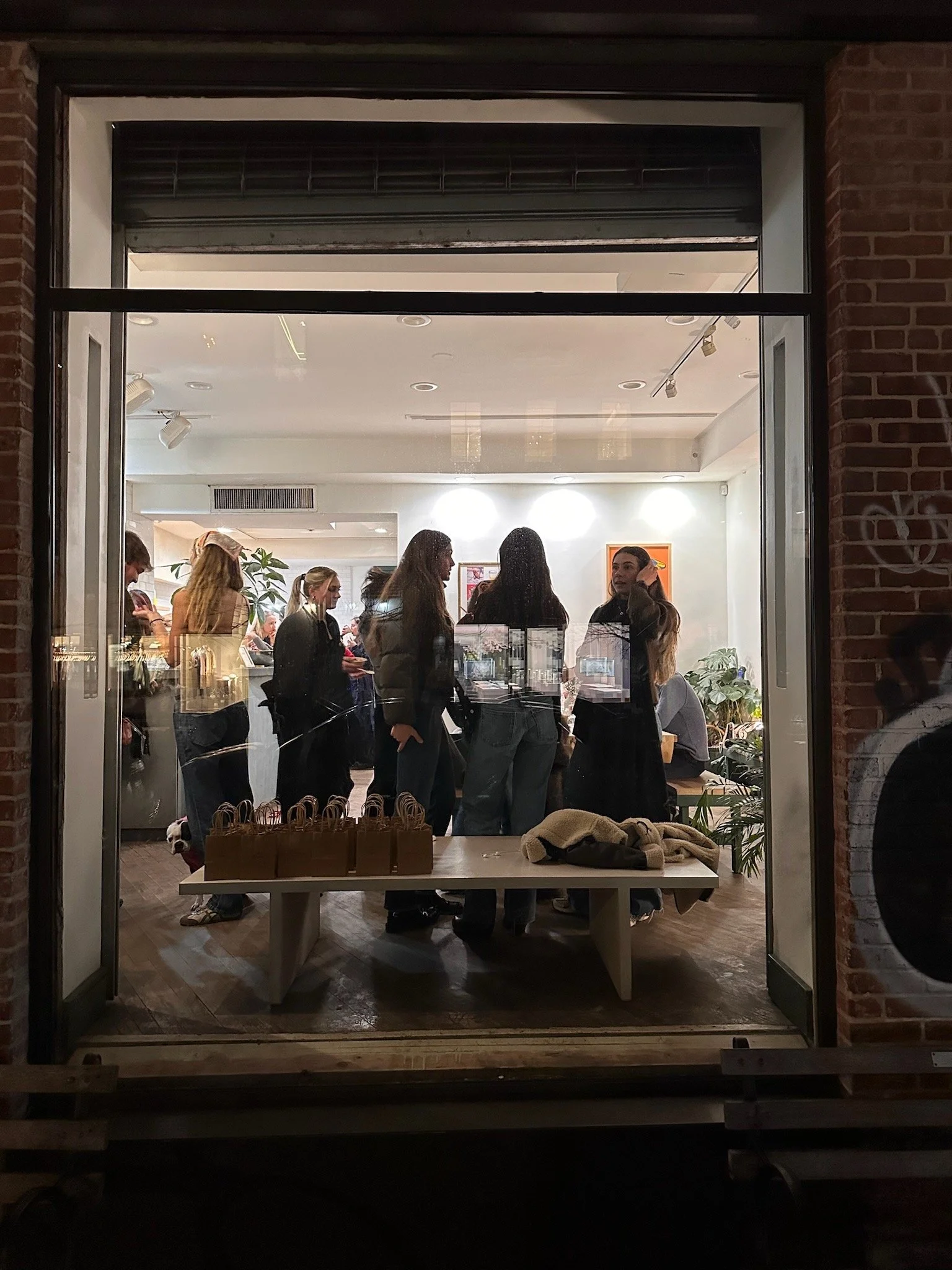 People inside a well-lit room viewed through a large window at night, with a table outside holding gift bags and a jacket.