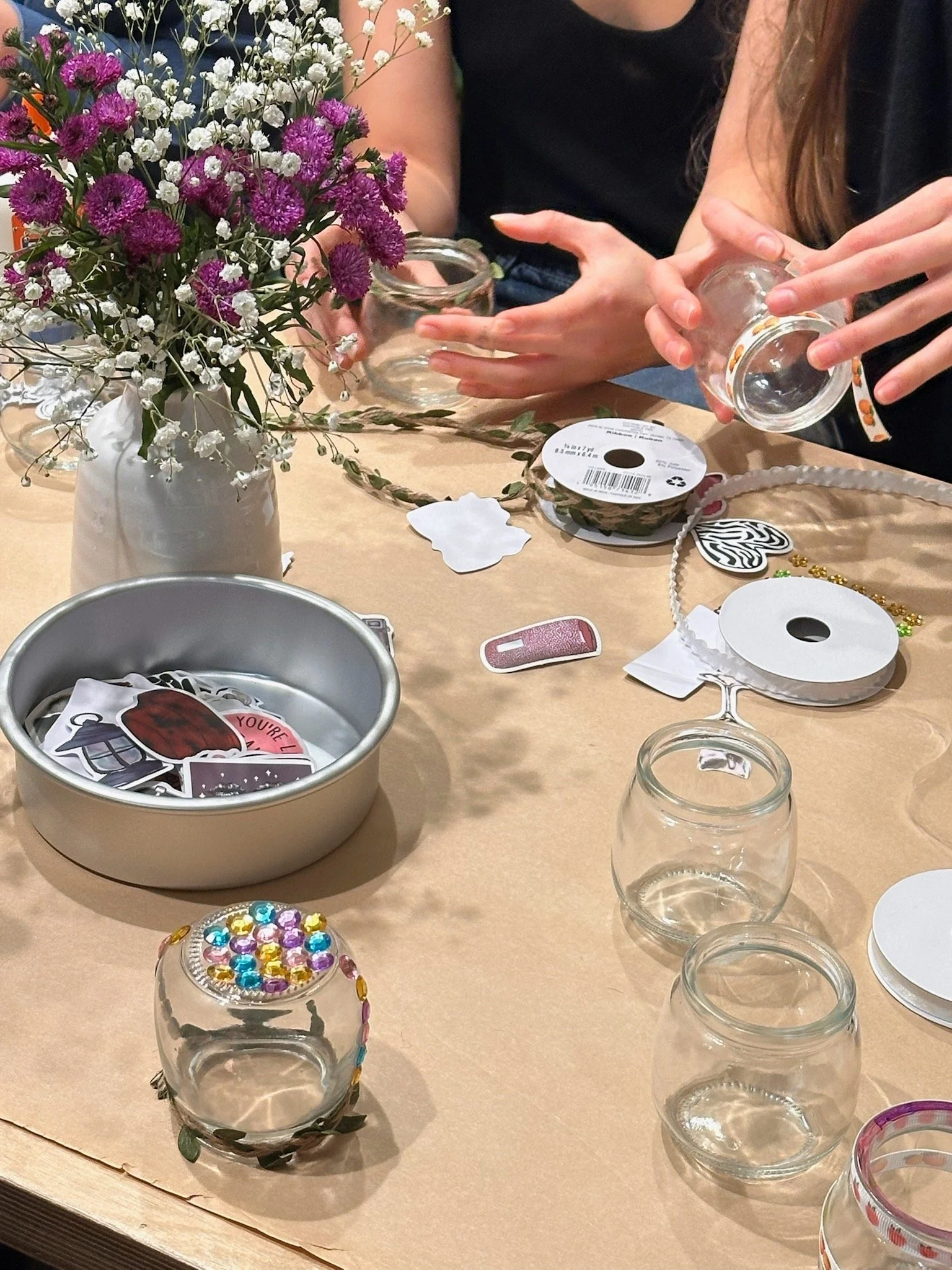 A table with crafting supplies including glass jars, decorative stickers, ribbons, and a vase of purple and white flowers, with two people arranging items on the table.