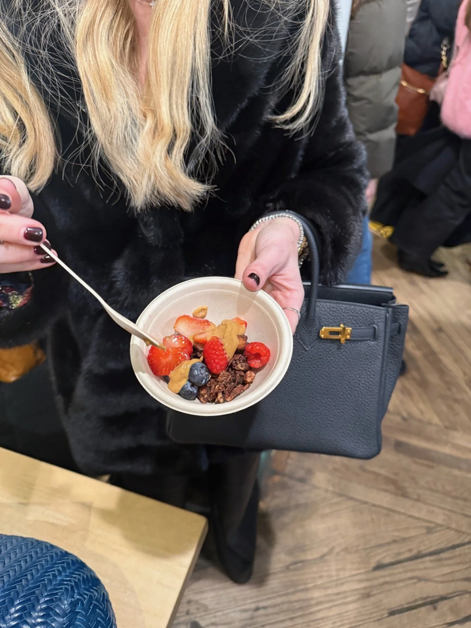 Person holding a bowl of yogurt with strawberries, blueberries, raspberries, banana slices, and granola, using a small spoon.