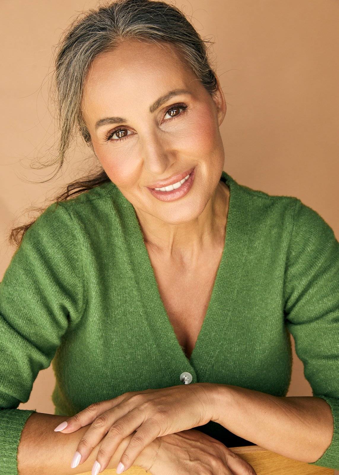 A smiling middle-aged woman with brown eyes and gray hair tied back, wearing a green sweater, sitting with her hands clasped on a wooden surface against a peach-colored background.