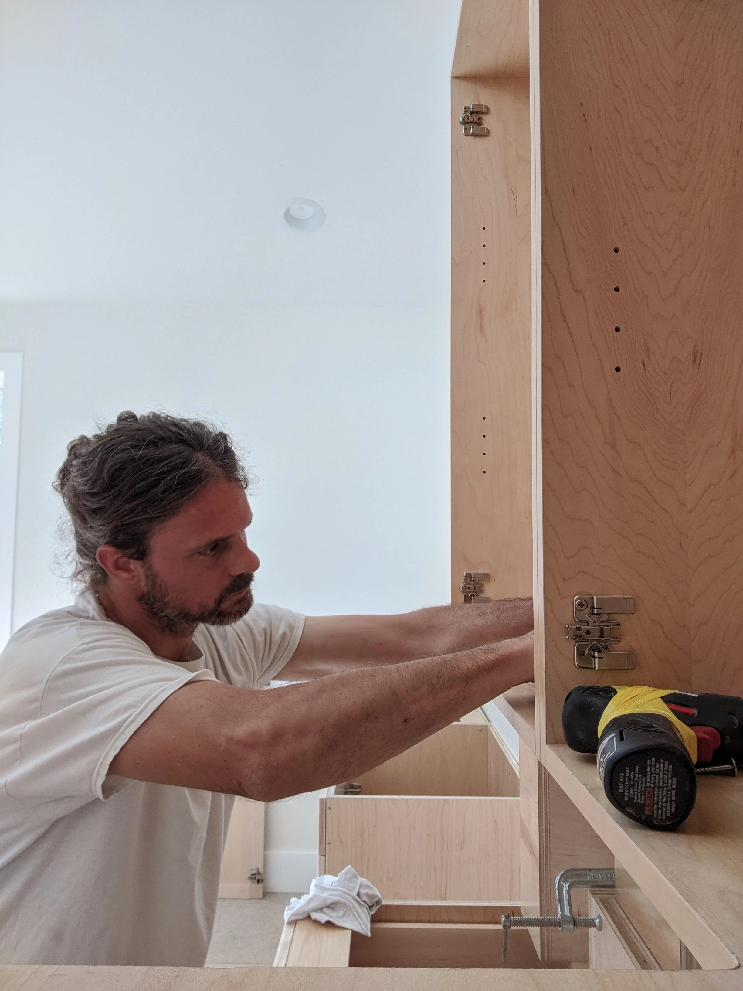 A man with curly hair and a beard, wearing a white t-shirt, is installing a wooden cabinet door with hinges in a light-colored room. He is using a power drill on the right side of the image, and wood shavings are visible on a towel on the countertop.