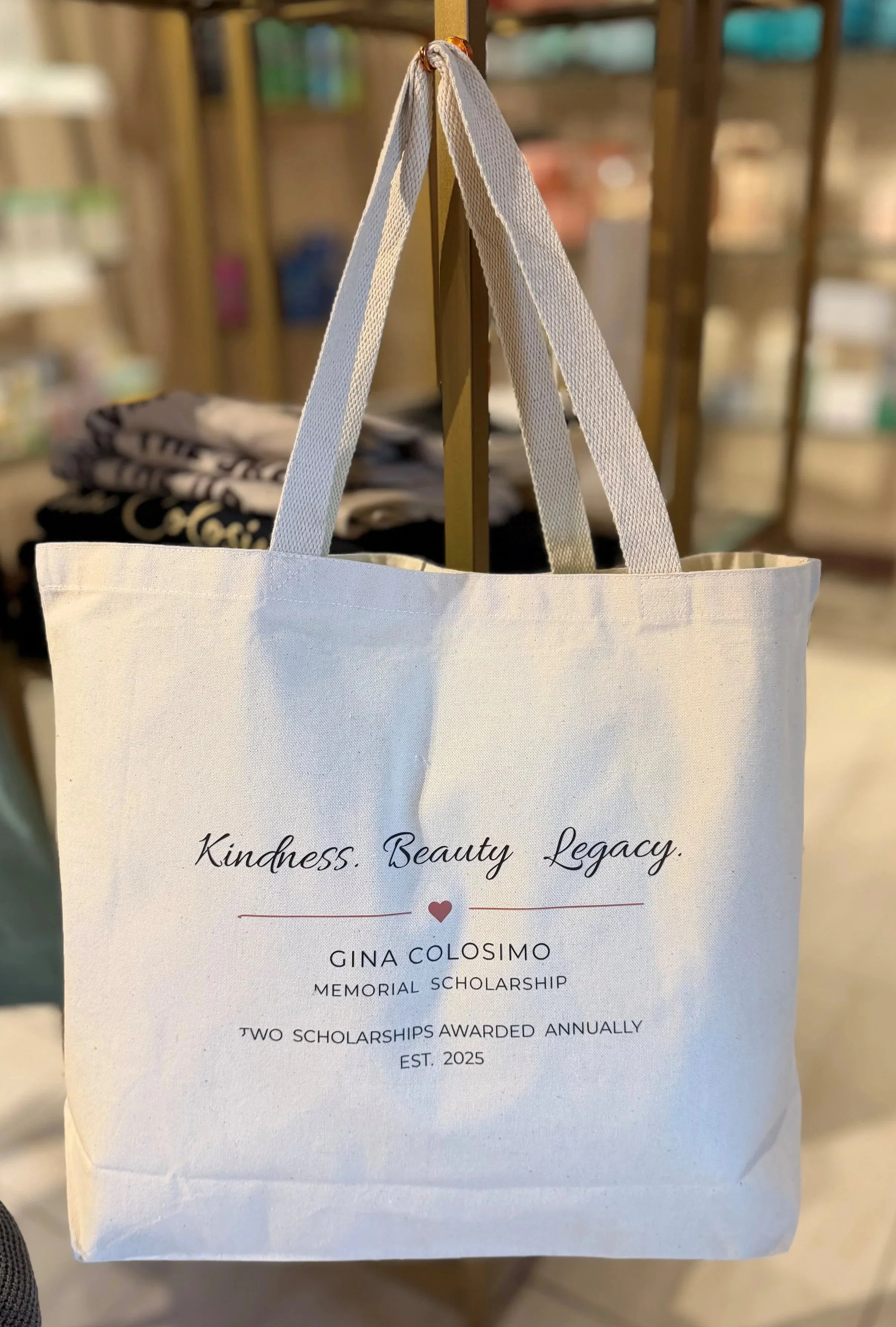 White tote bag with black cursive text reading "Kindness. Beauty. Legacy." and additional text about Gina Colosimo memorial scholarship printed on it.