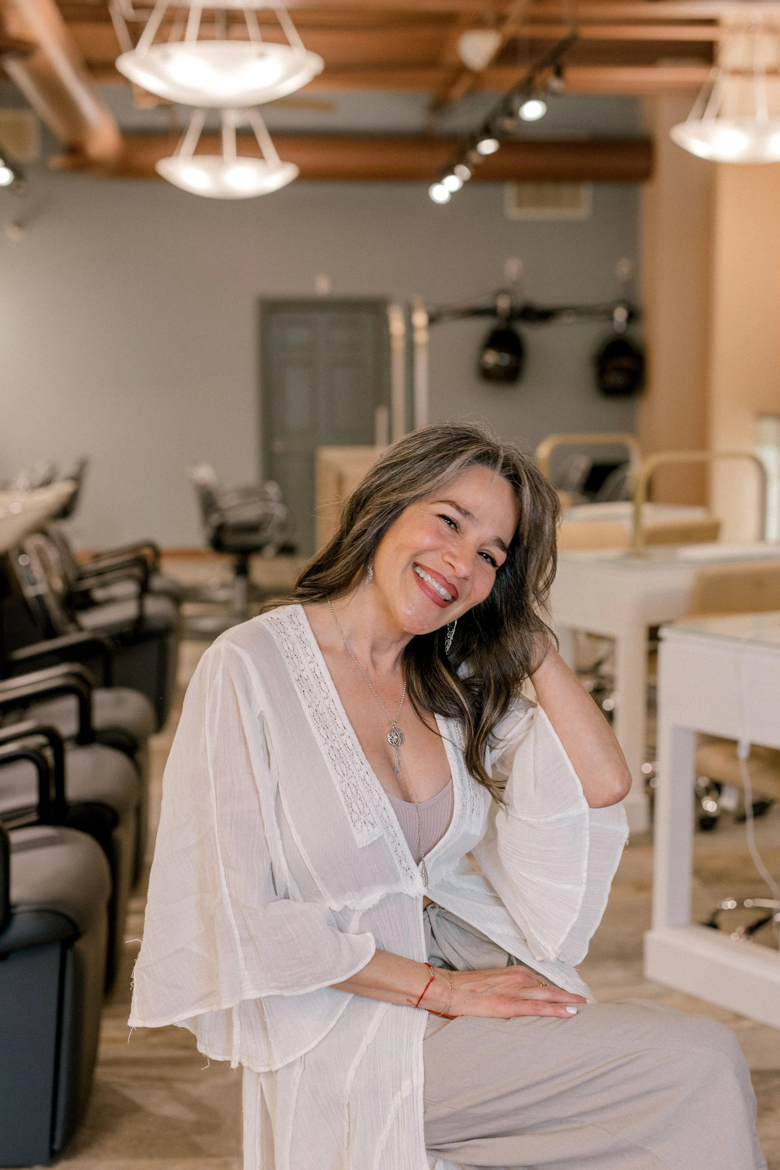A woman with brown hair and light skin sitting on a beige chair, wearing a white shirt and blue jeans, smiling at the camera, with a plain white background.