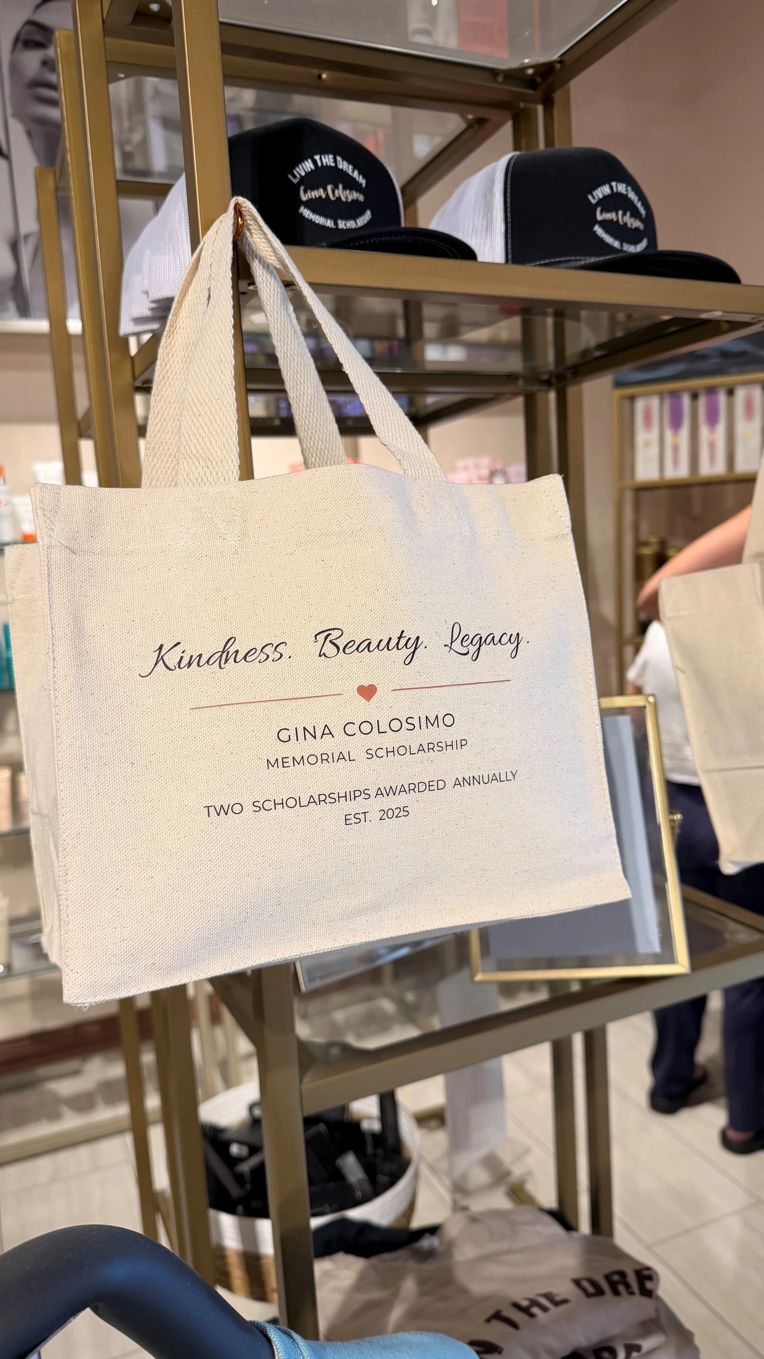 A beige tote bag with the text 'Kindness, Beauty, Legacy' and details about Gina Colosimo Memorial Scholarship, established in 2025, and awarded annually, hanging on a gold display stand in a retail or event space with hats and framed photos in the background.