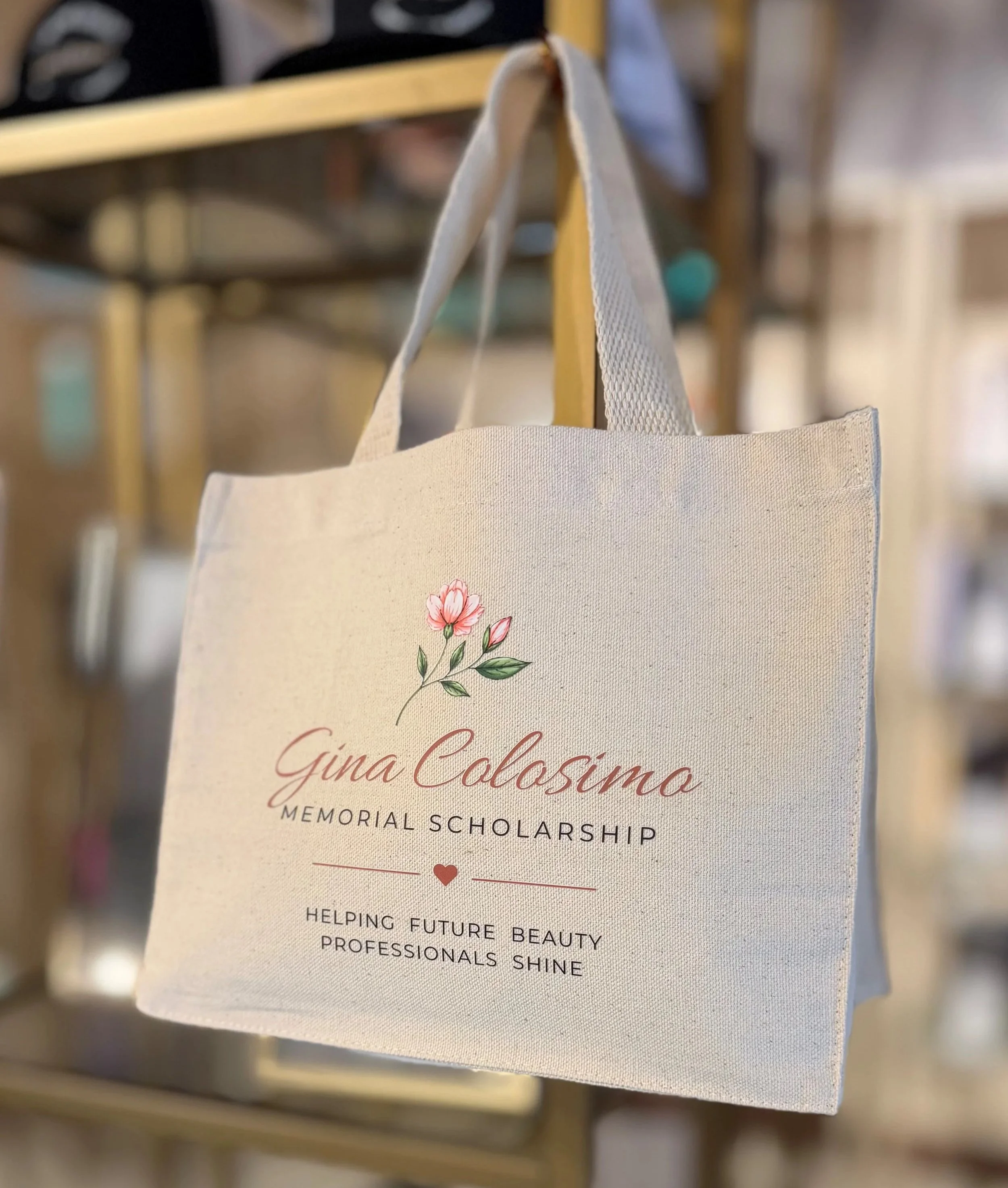 A white tote bag with pink flowers and text commemorating Gina Colosimo, a Memorial Scholarship supporting future beauty professionals.