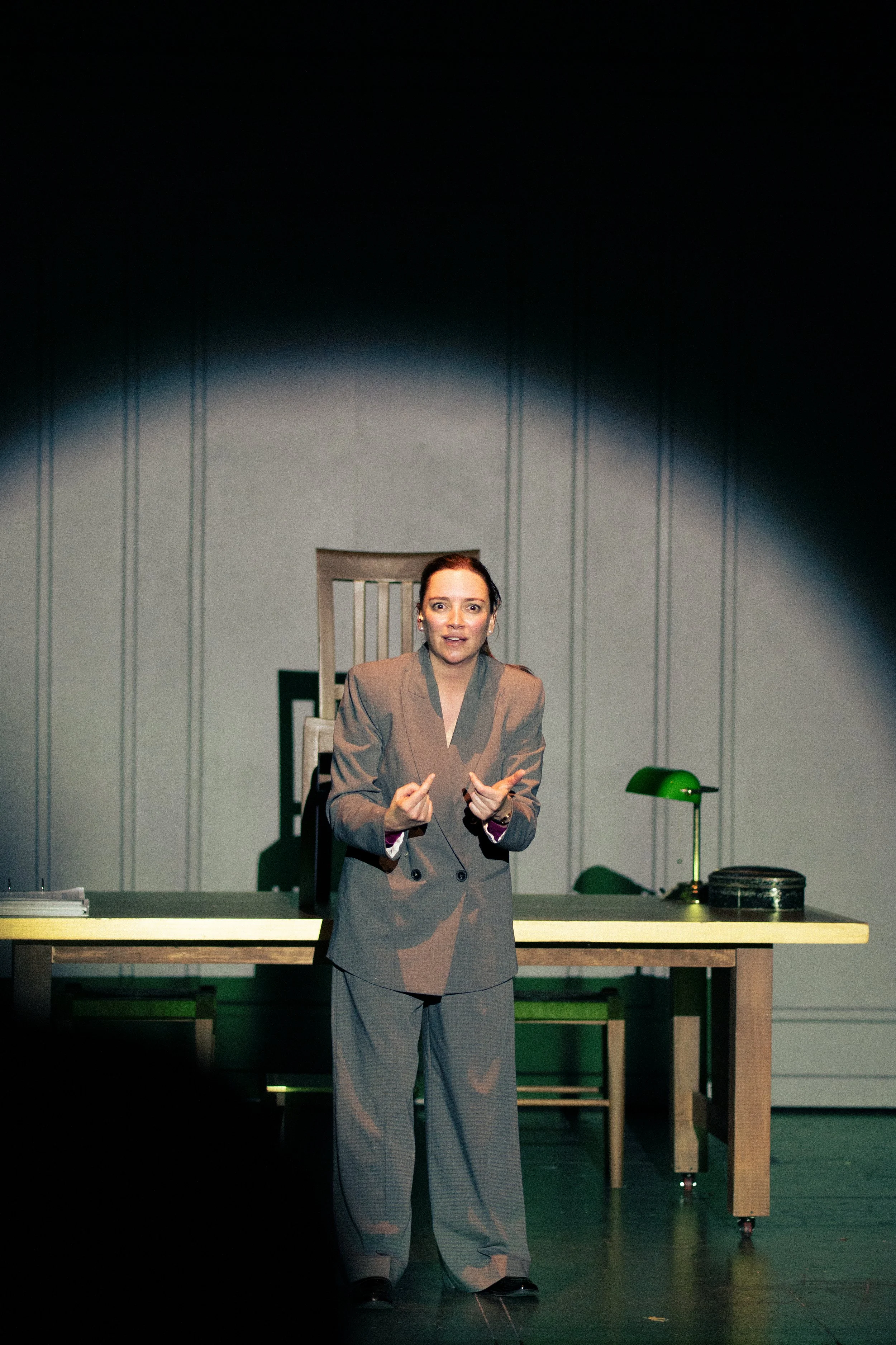 Una mujer vestida con un saco gris y pantalones a cuadros, gesticulando con las manos, en un escenario con una mesa y una lámpara verde.