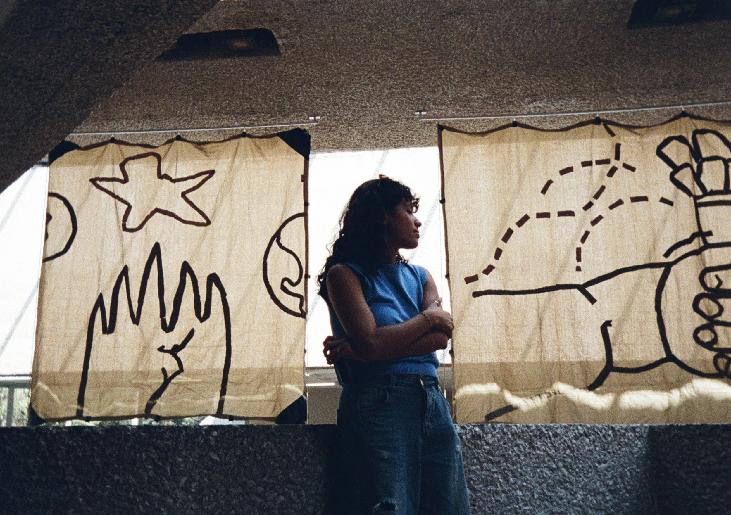 Una joven en un espacio cerrado, con fondos de cortinas de tela beige con dibujos de líneas negras de un planeta, una estrella, un delfín y una mano.