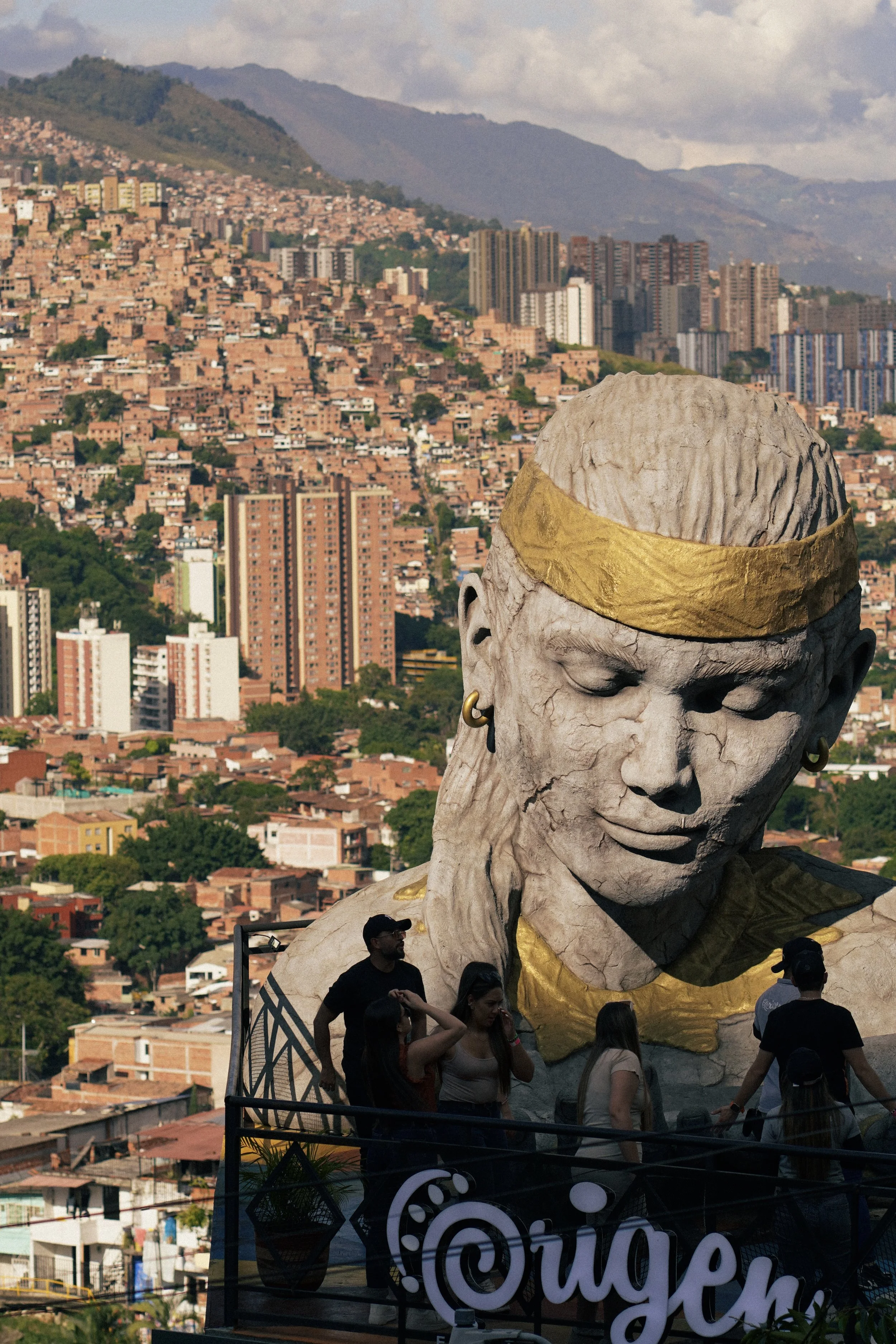 Monumento en forma de rostro de mujer con accesorios dorados y aretes, con fondo urbano de colinas y edificios en una ciudad.