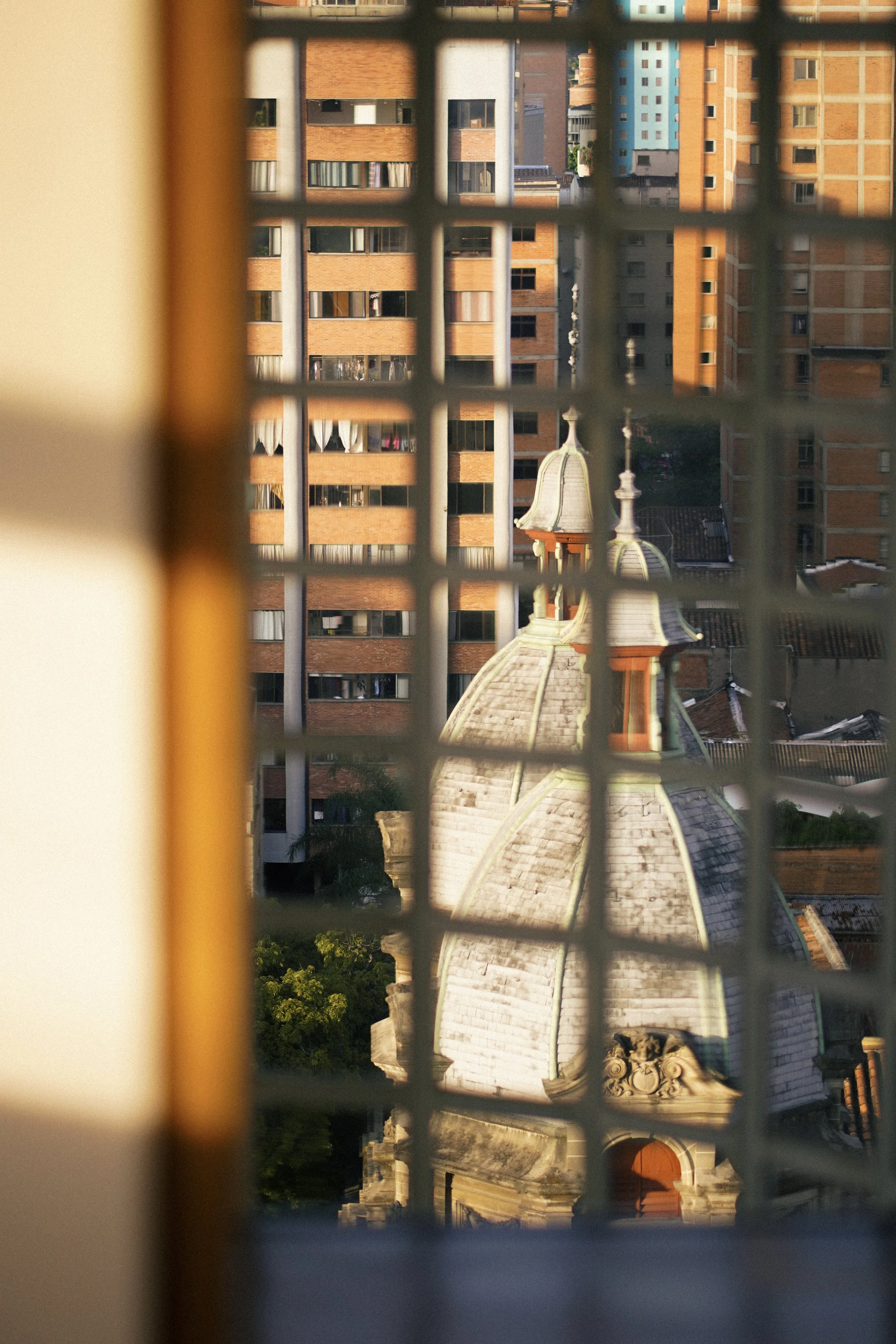 Vista de una iglesia antigua con cúpula y campanario, vista a través de una ventana con rejas, en medio de edificios modernos altos y de apartamentos en Bogotá, Colombia.