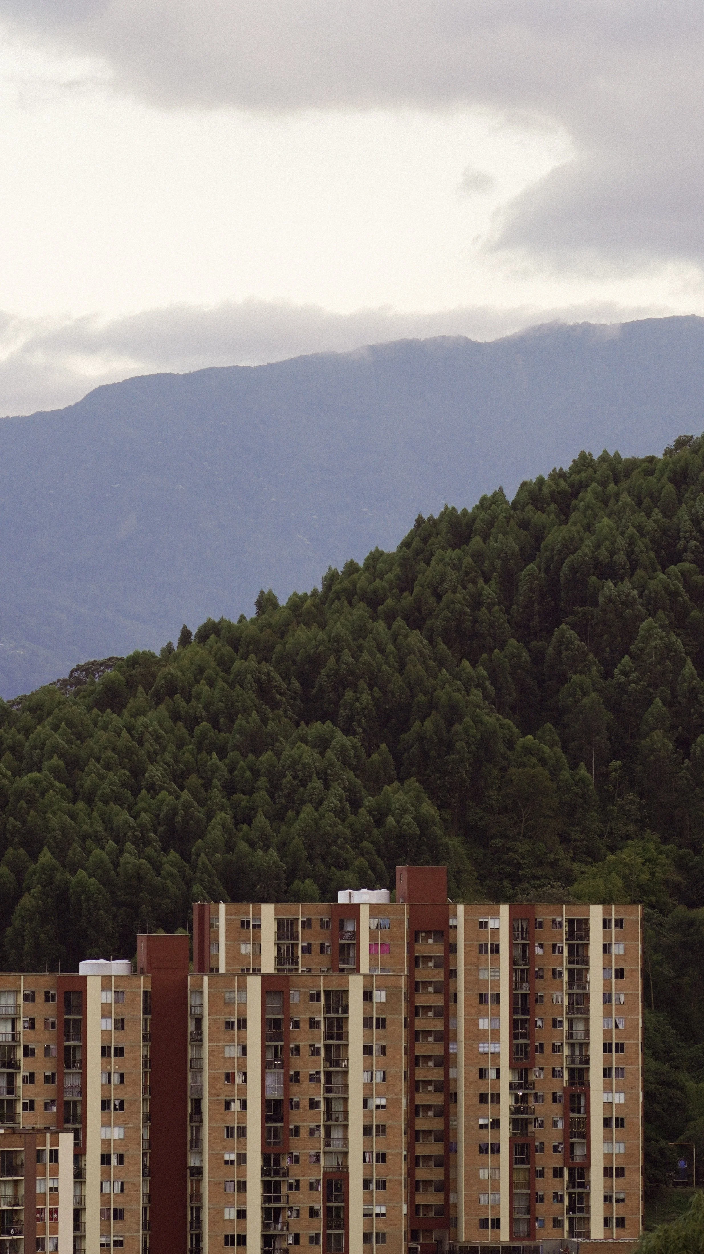 Edificio de apartamentos en primer plano, con montañas verdes y nubes en el fondo, un día nublado.
