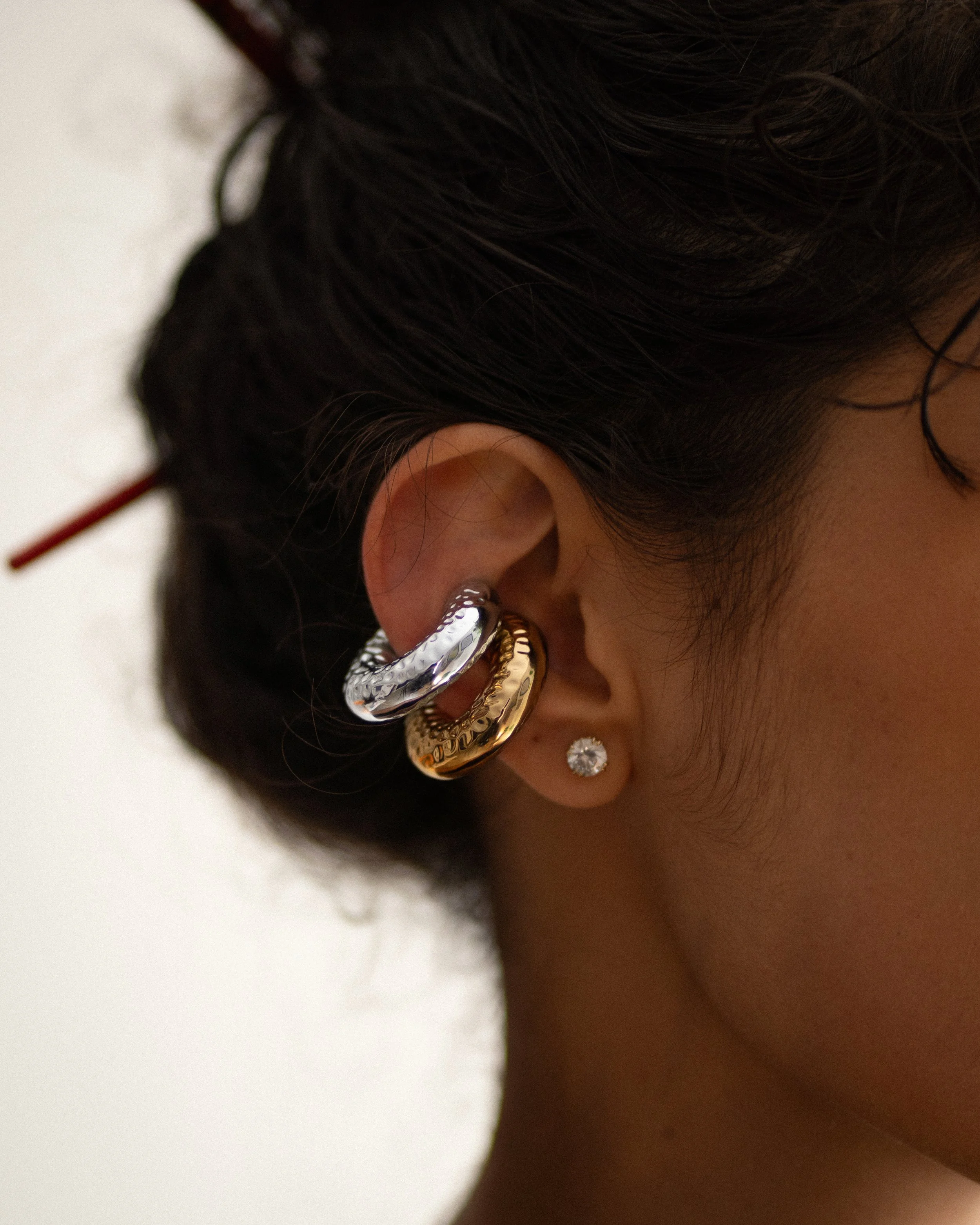 Primer plano del oído de una persona con tres pendientes grandes y una pequeña en forma de diamante en el lóbulo, el cabello oscuro y rizado parcialmente recogido.