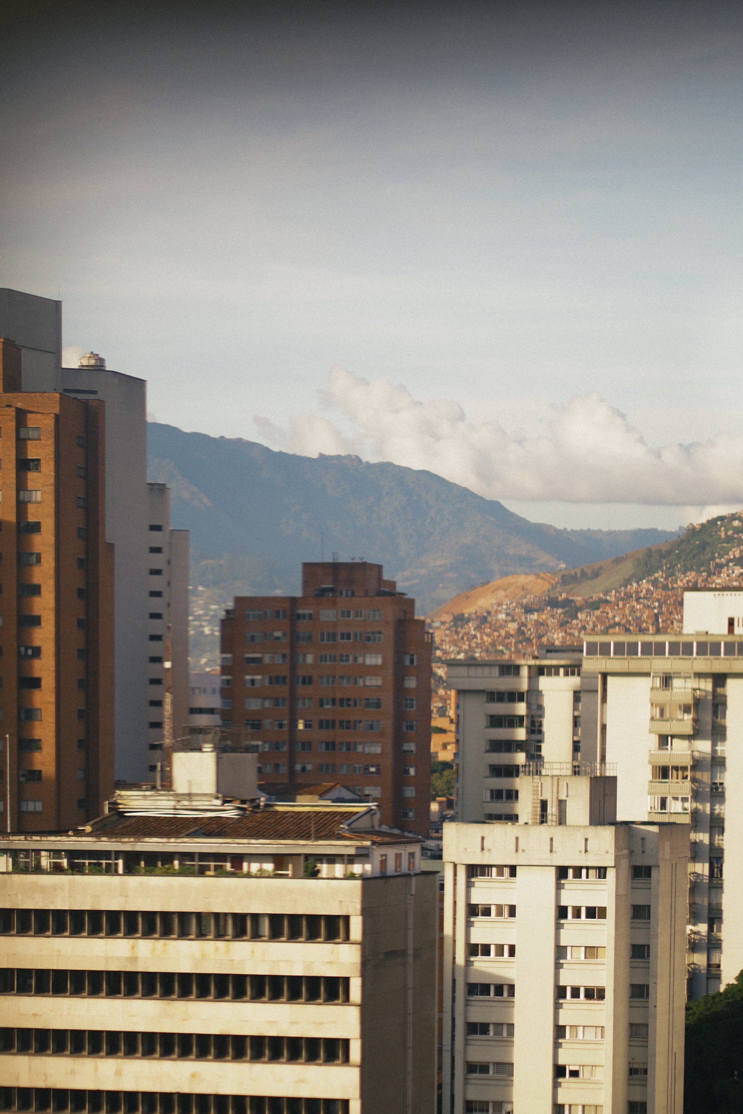 Vista de una ciudad con edificios altos y montañas al fondo, cielo despejado con algunas nubes.