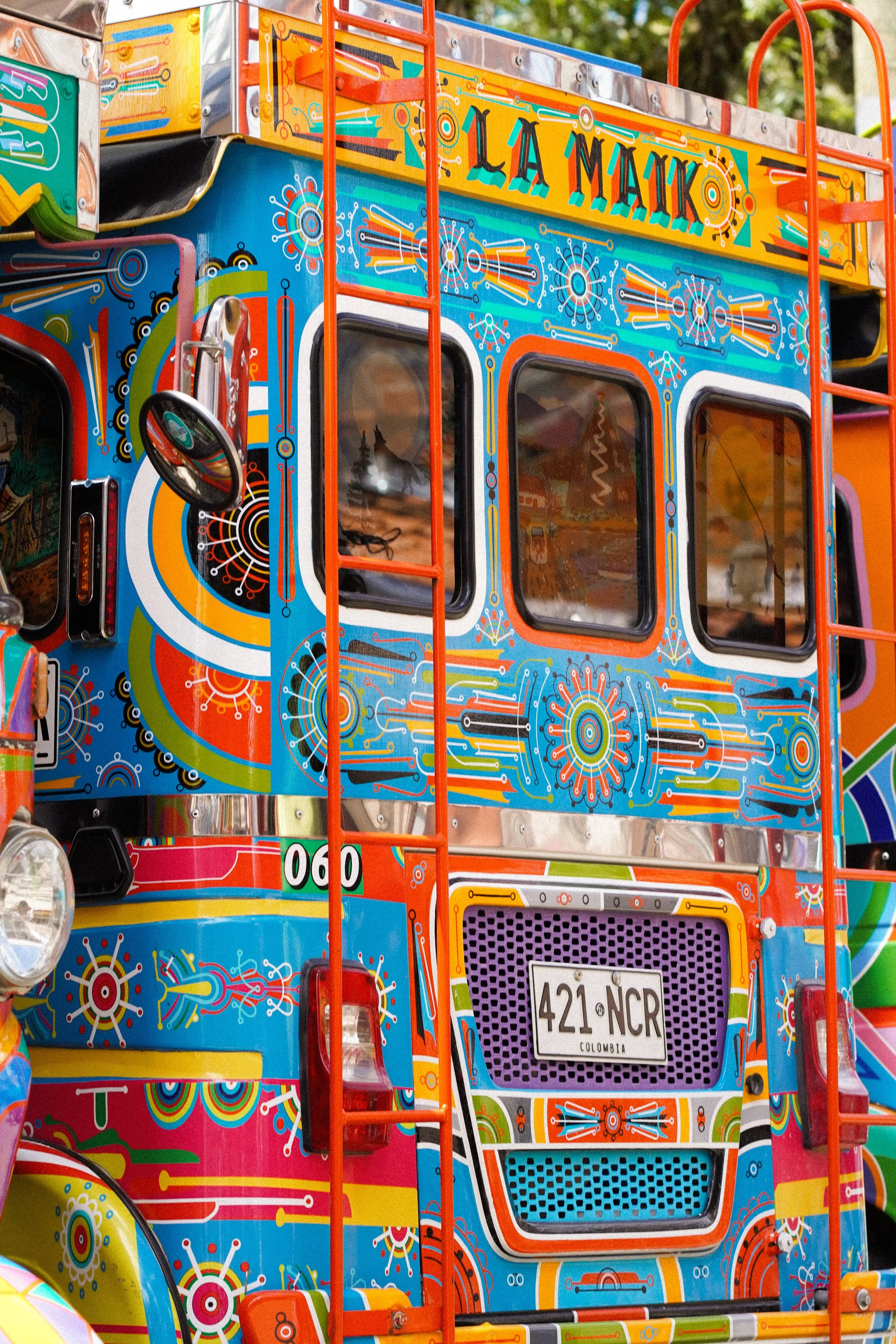 Parte frontal de un vehículo decorado con colores vivos y patrones geométricos, con ventanas, espejos, luces y una matrícula que dice '421 NCR' de Colombia.