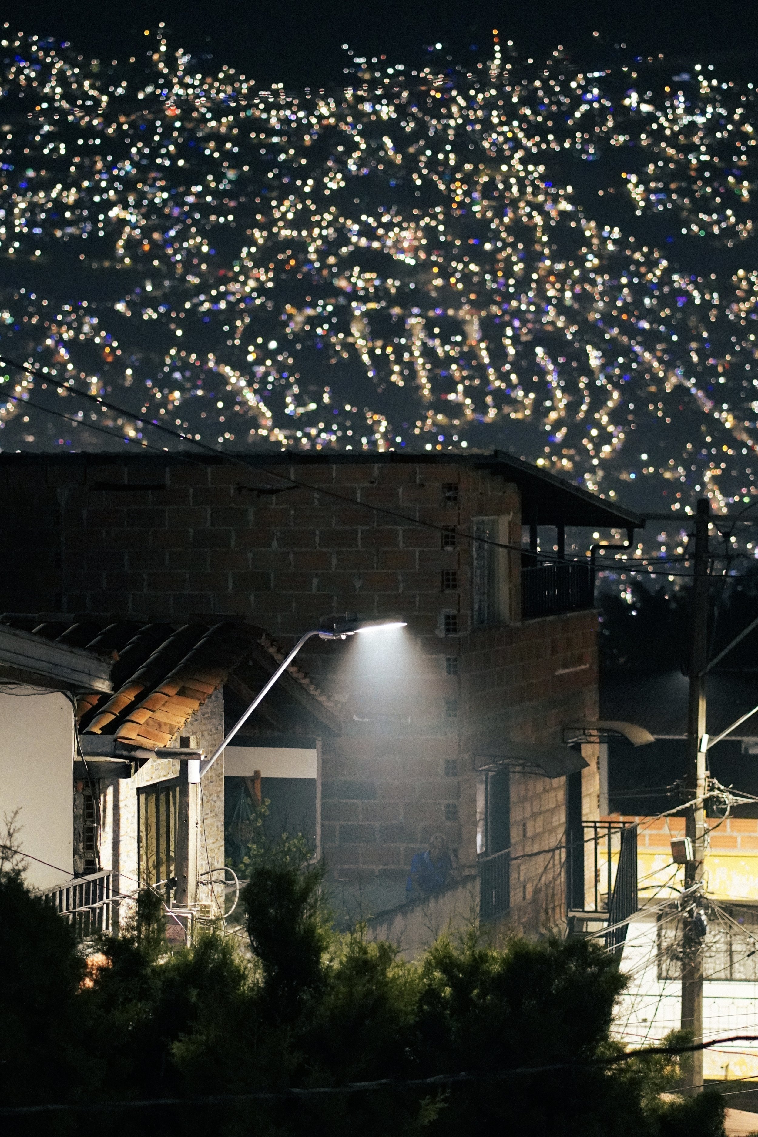 Vista nocturna de un barrio con casas de ladrillo y algunas plantas, iluminado por una lámpara en la calle, con un fondo de la ciudad con muchas luces difusas en las colinas.