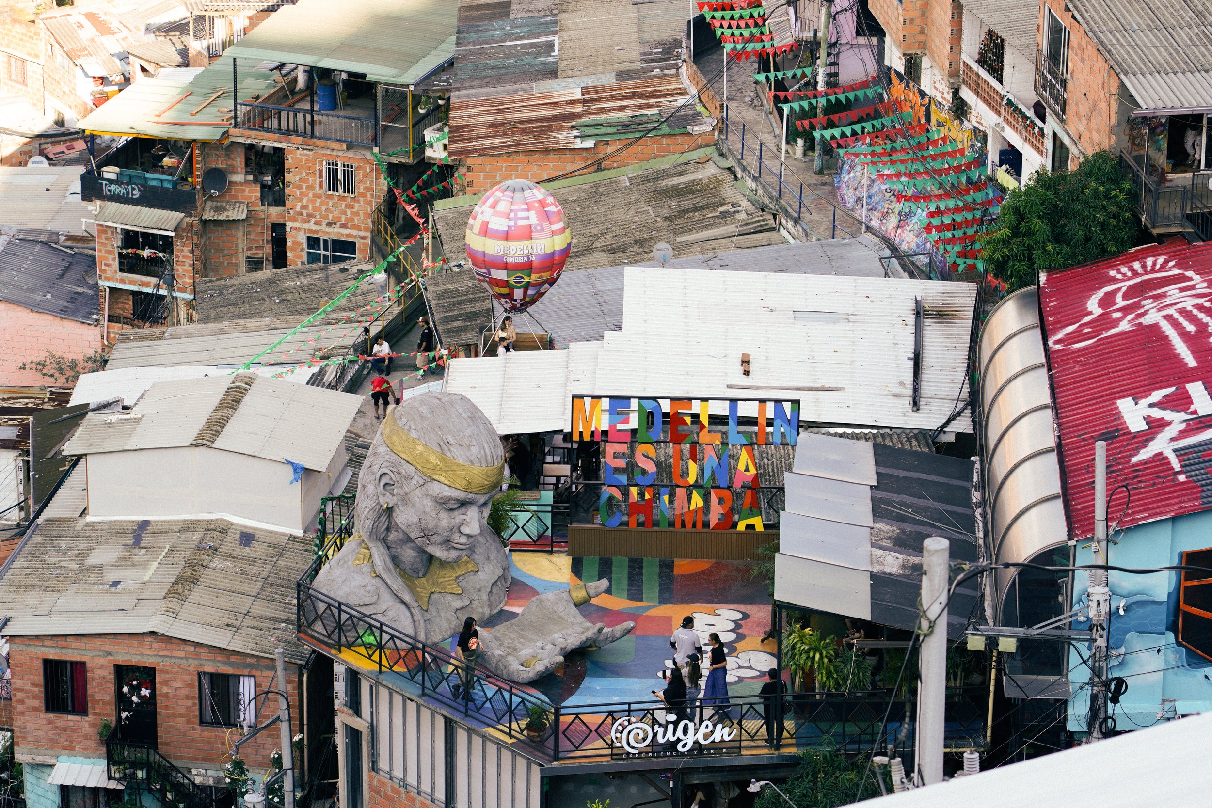 Vista aérea de un vecindario con casas de techos de lámina, algunos con paredes de ladrillo y otros pintadas. En el centro se observa una plaza con una escultura grande de una mujer, un globo aerostático decorativo y una señal colorida que dice 'MEDE
