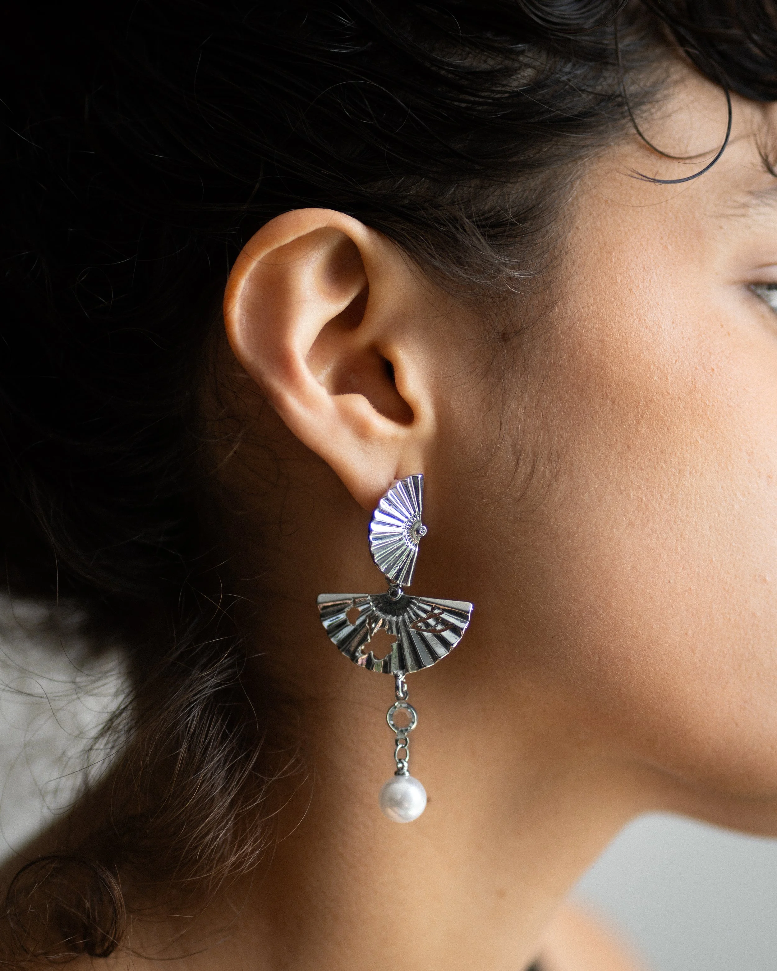 Un close-up de la oreja de una mujer con un pendiente de diseño metálico en forma de abanico, con un colgante de perla en la parte inferior.