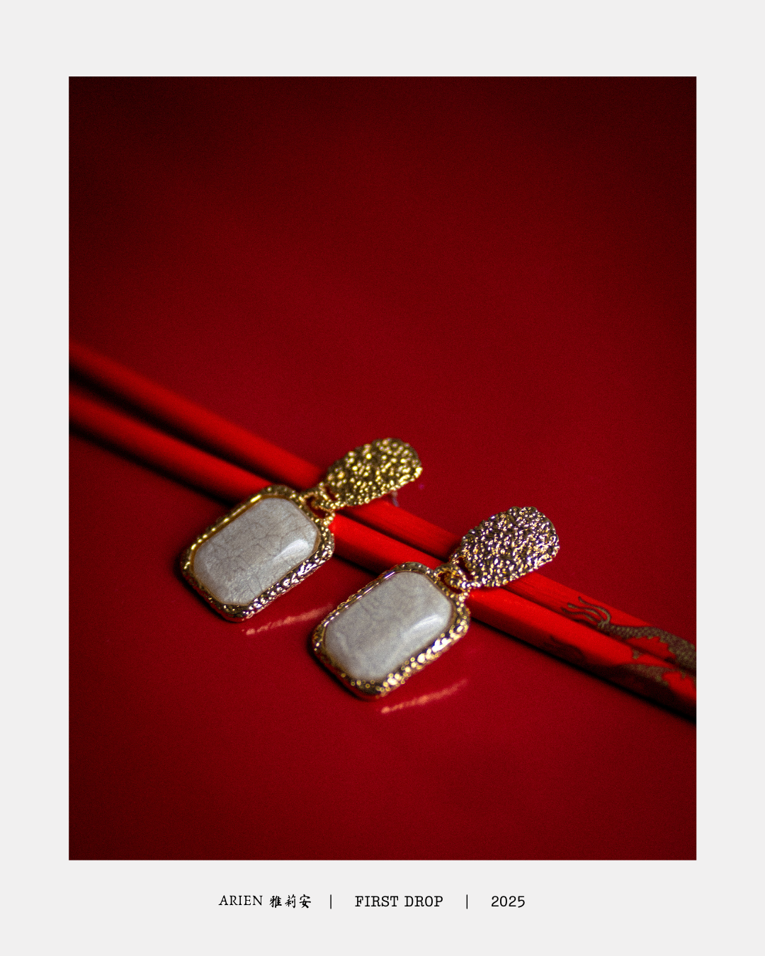 Aretes dorados con piedra blanca en un fondo rojo, etiquetados como ARIEN, FIRST DROP, 2025.