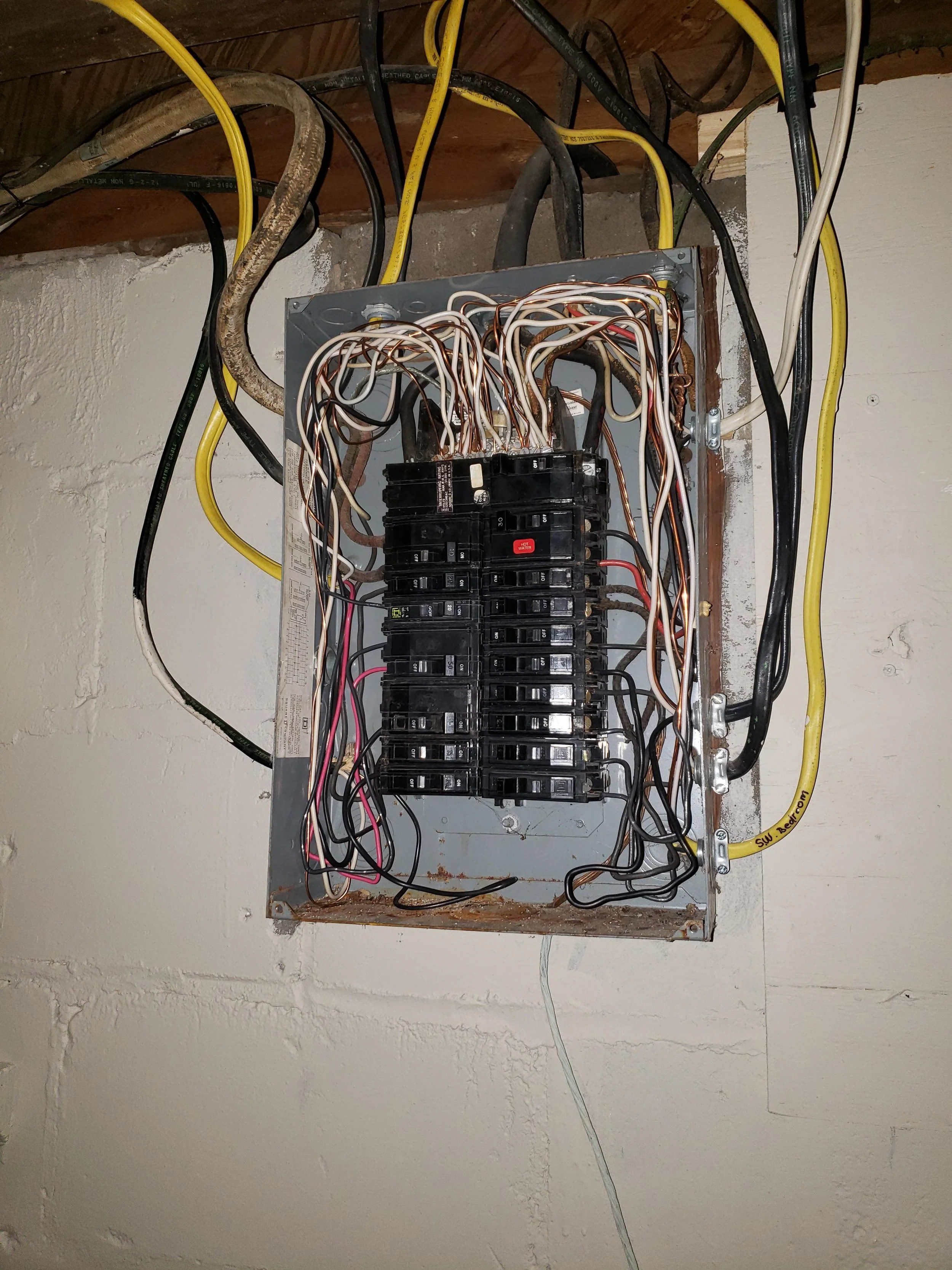 Electrical breaker box with open cover, showing circuit breakers and connected wires of various colors, mounted on a wall with exposed wiring.