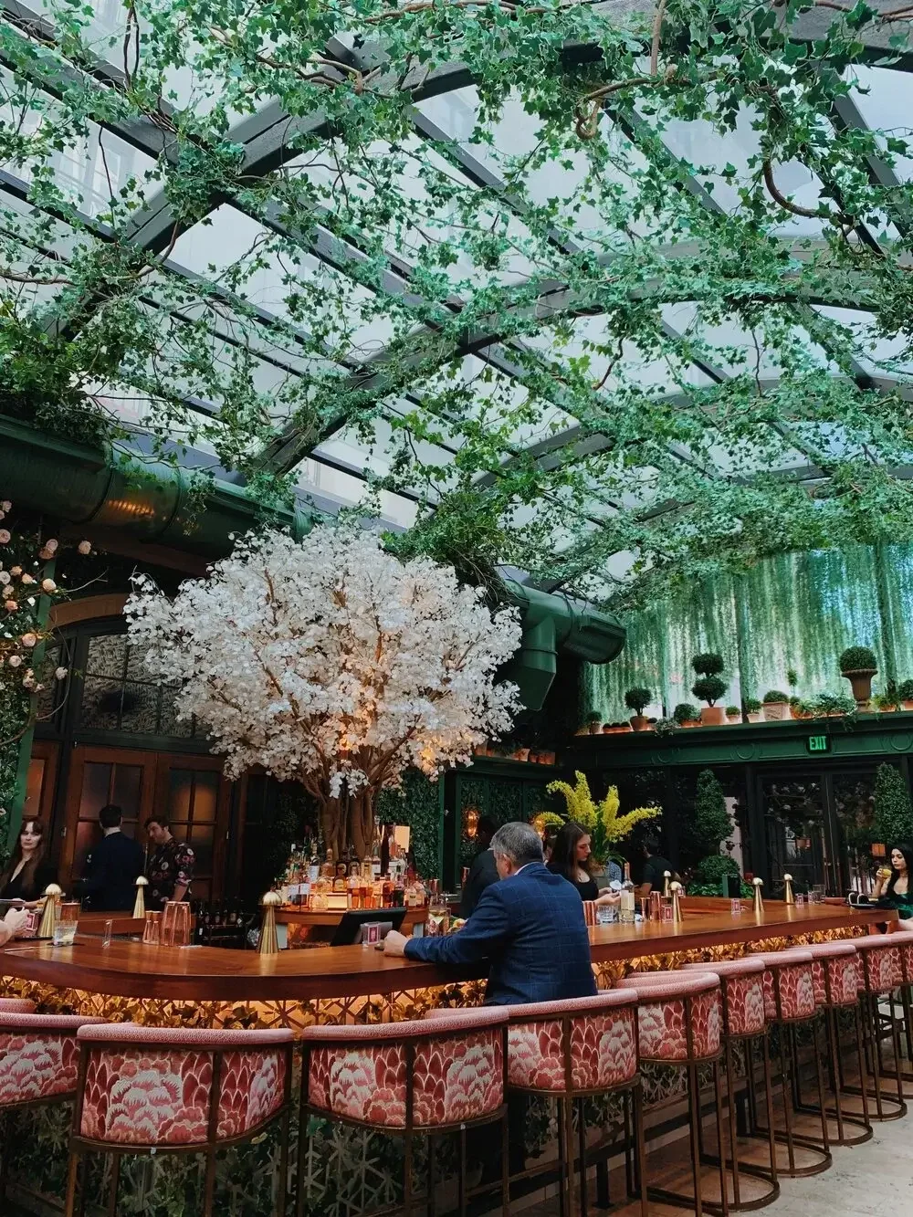 An elegant indoor bar featuring a stunning artificial cherry blossom tree at the center, creating a serene focal point within the space. A curved wooden bar counter with red patterned stools surrounds it, where guests are seated enjoying drinks and c