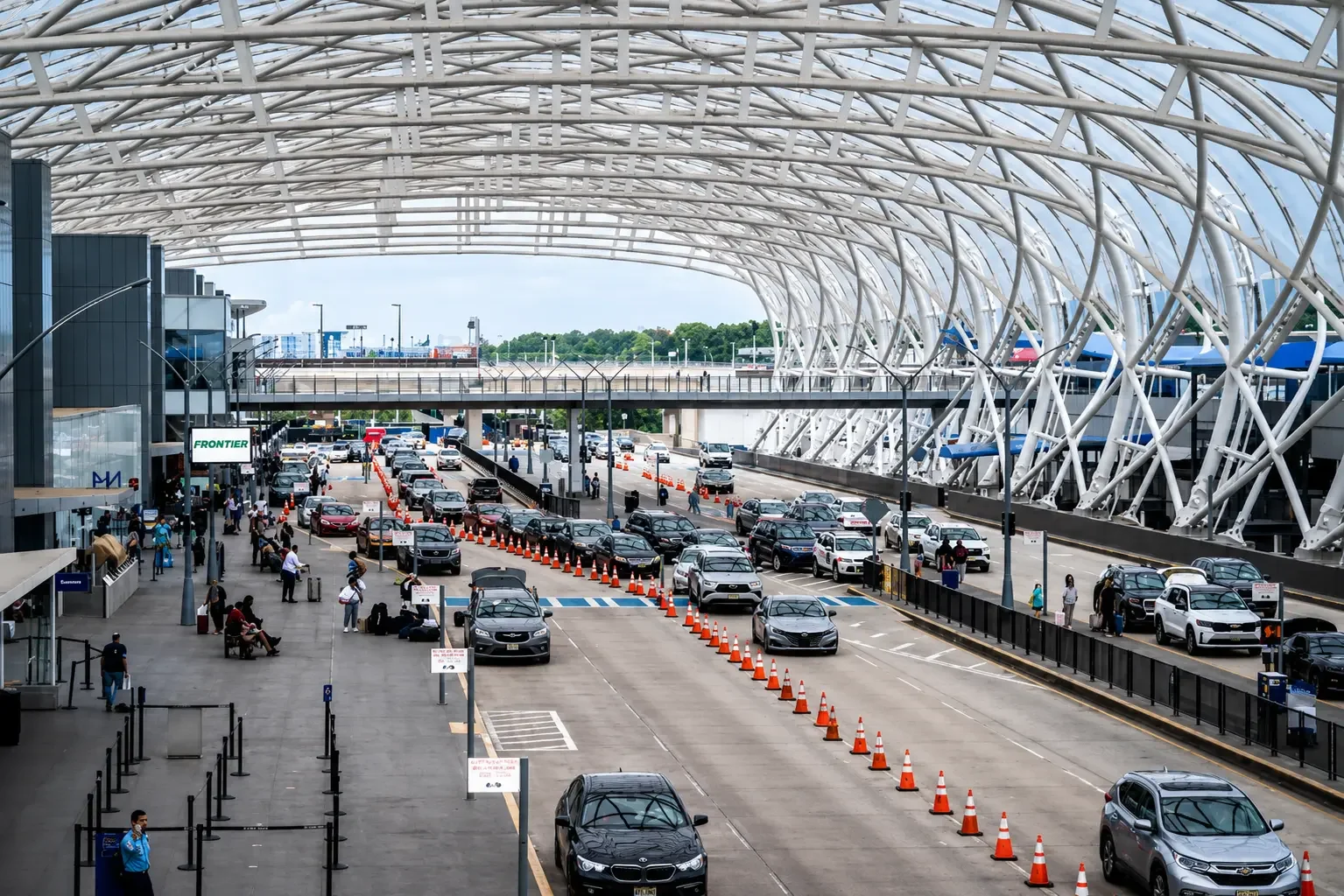 Vehicles lined up in a modern airport terminal with a distinctive white, curved, lattice-like roof structure. Passengers with luggage wait near the entrance, and signs are visible.