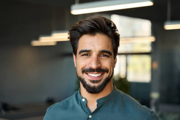 A man with dark hair and a beard smiling in an indoor setting with large windows and modern lighting fixtures.