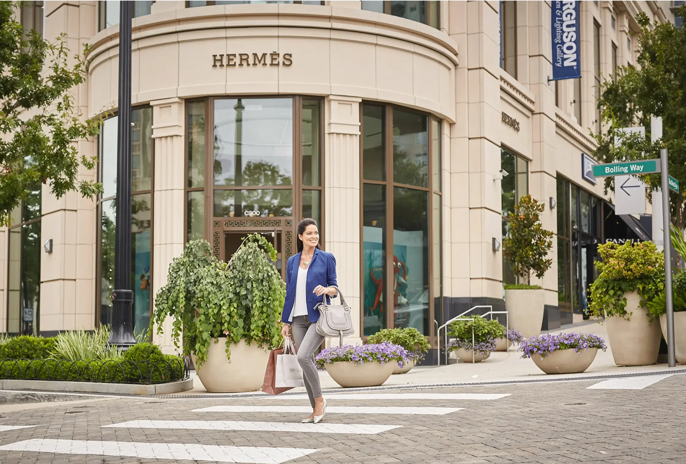 A woman in business attire walks confidently across a city street carrying shopping bags, passing in front of an elegant luxury boutique with large glass windows and refined architectural details. The storefront is framed with decorative elements and