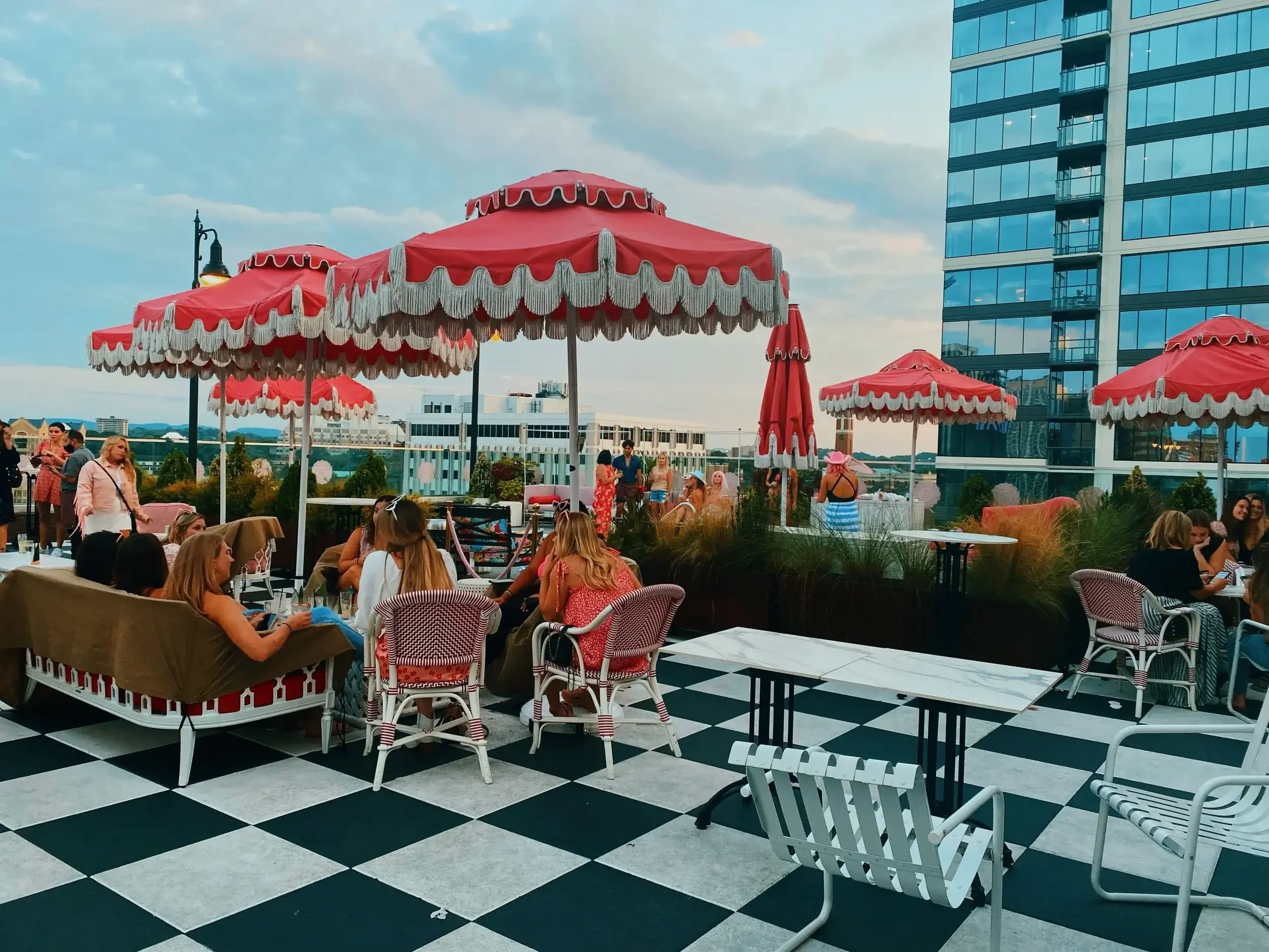 People dining on a stylish rooftop patio with red umbrellas and a black-and-white checkered floor, overlooking tall modern glass skyscrapers in a vibrant city skyline. The atmosphere reflects luxury urban living and premium lifestyle experiences. Bla