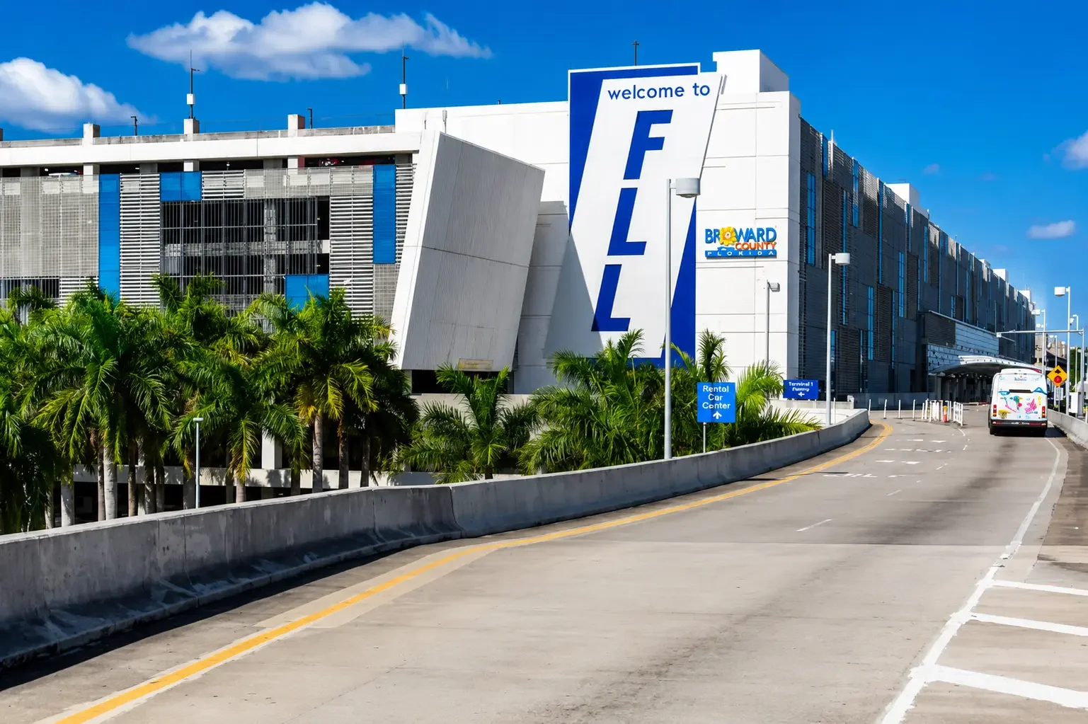 The image shows a large parking garage with a sign saying 'welcome to FLL' for Fort Lauderdale-Hollywood International Airport, with a blue sky and some clouds in the background and lush green palm trees in front.