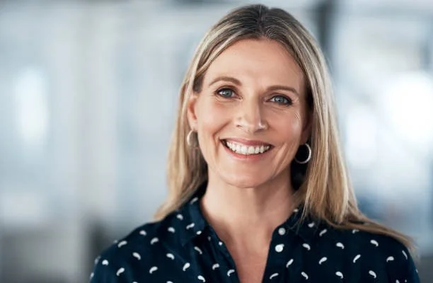 A smiling woman with shoulder-length blonde hair, wearing hoop earrings and a navy blue blouse with white polka dots, standing in an office setting.