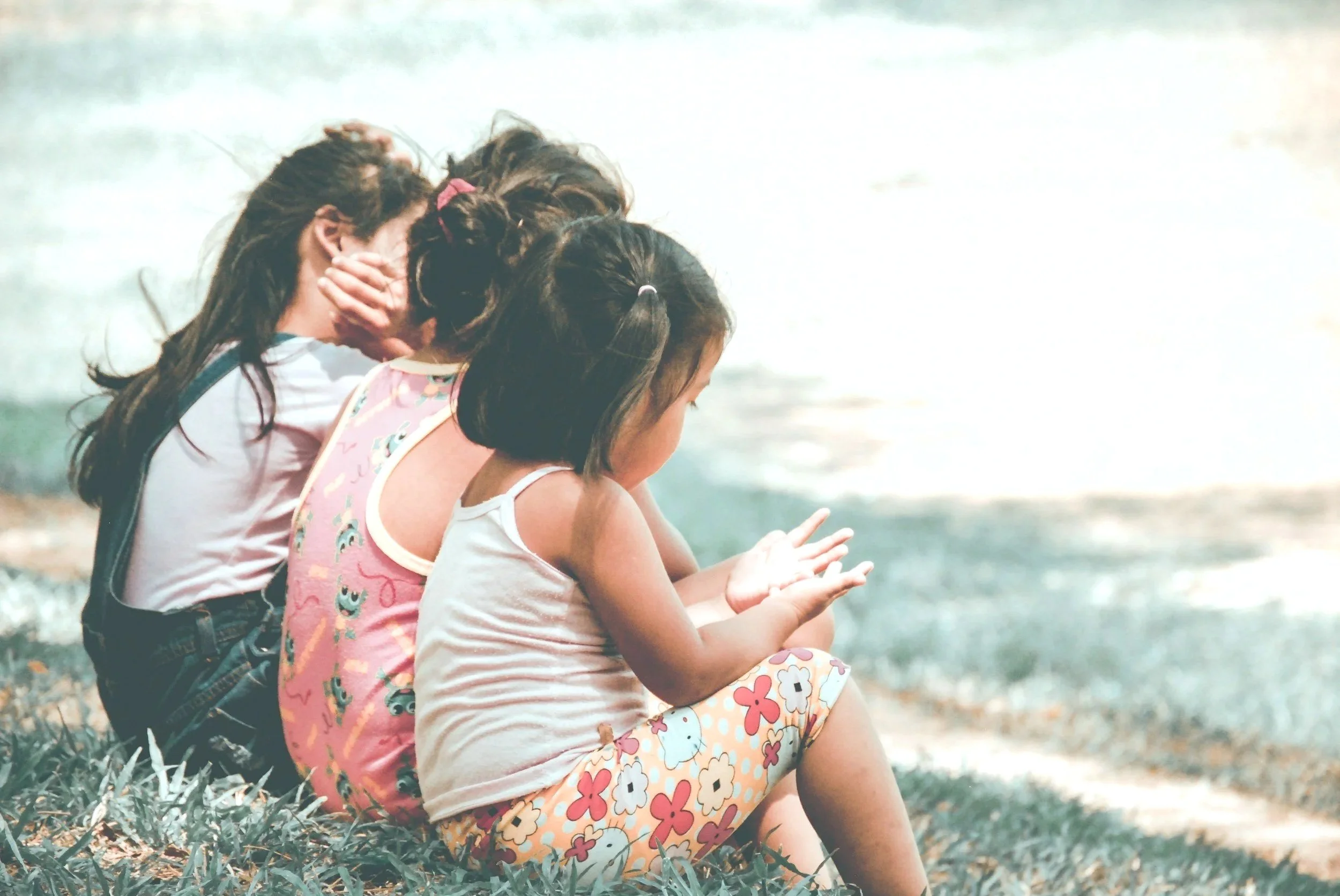 Three young girls sitting on grass near a body of water,bowing their heads and covering their faces, with the youngest looking at her open hands.