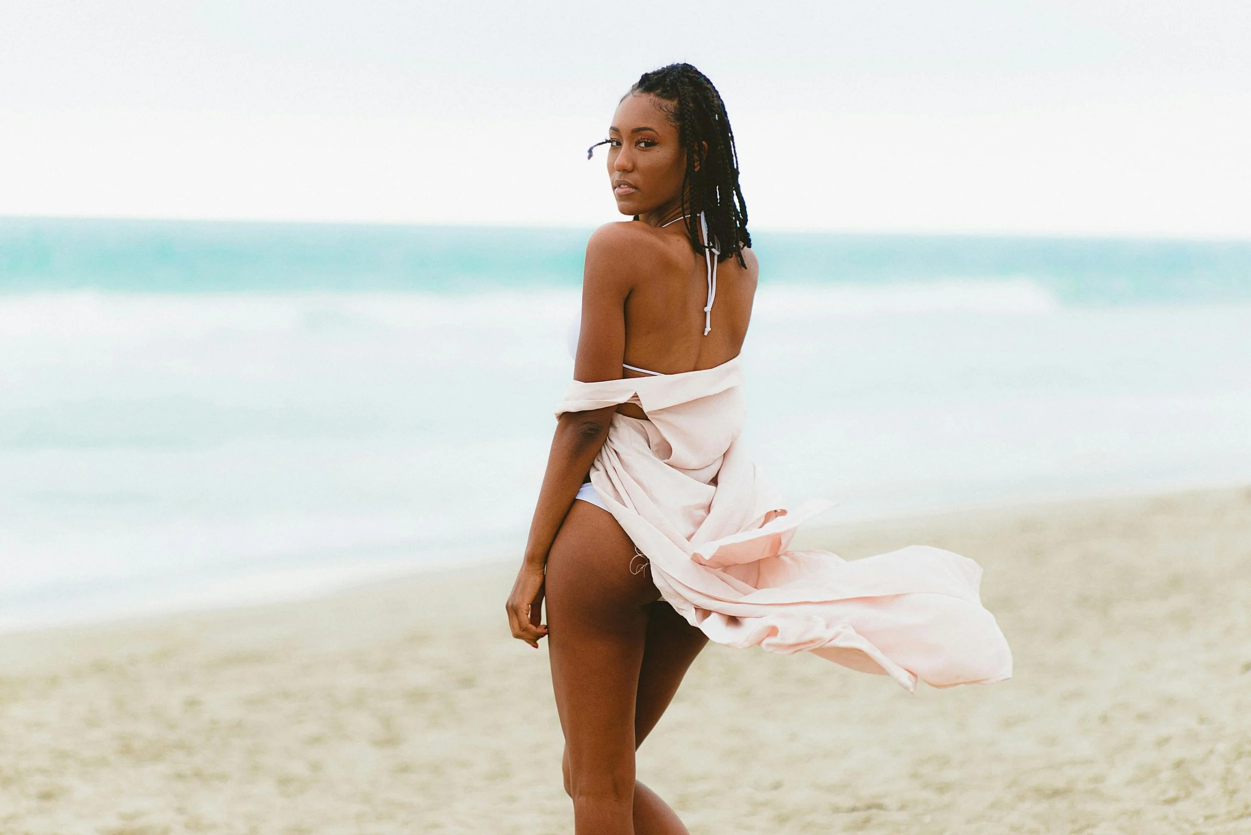 A woman with dark braided hair standing on the beach, wearing a light-colored cloth around her waist with the ocean in the background.