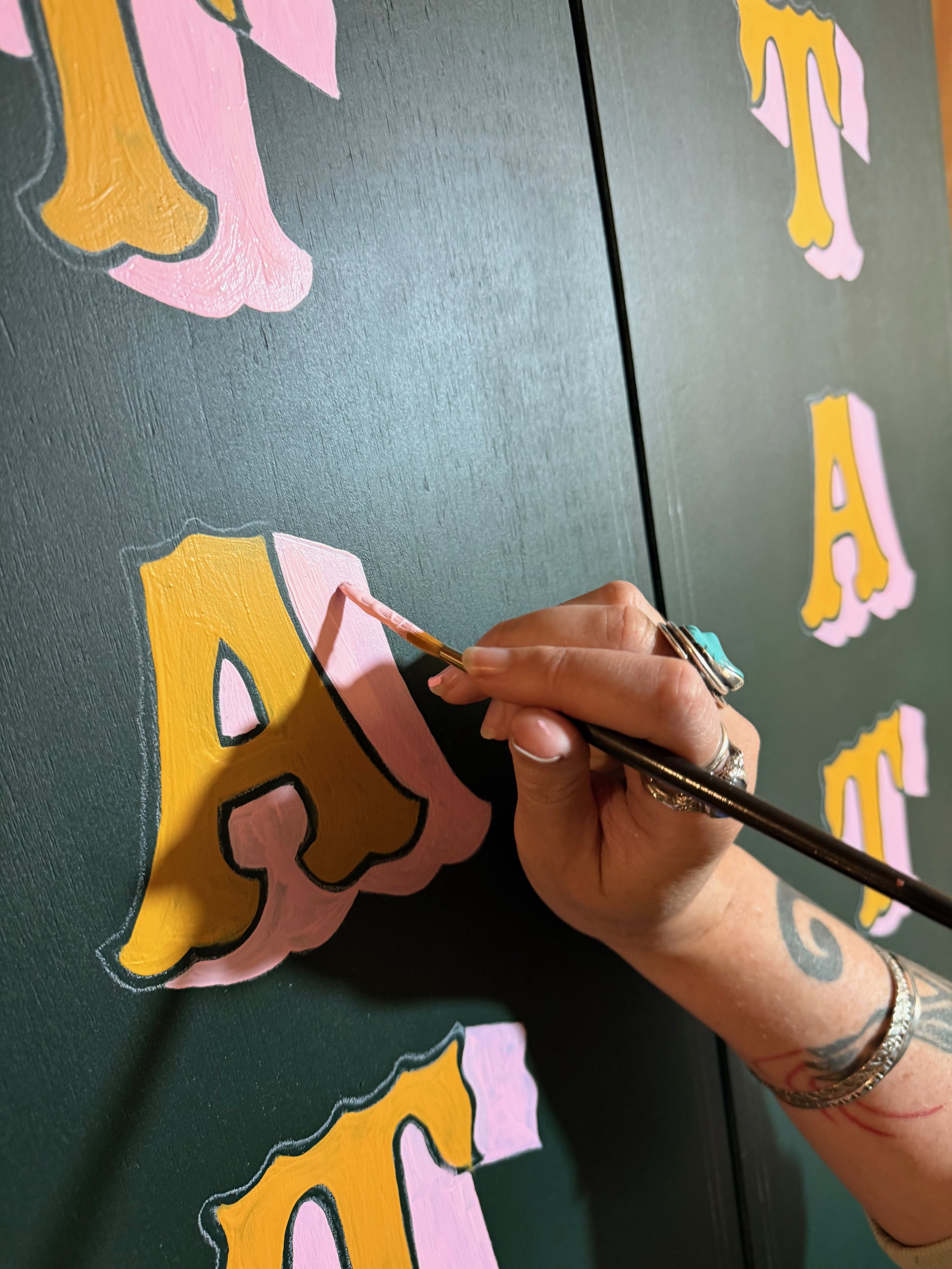 A person painting pink and yellow letters on a dark green wall.