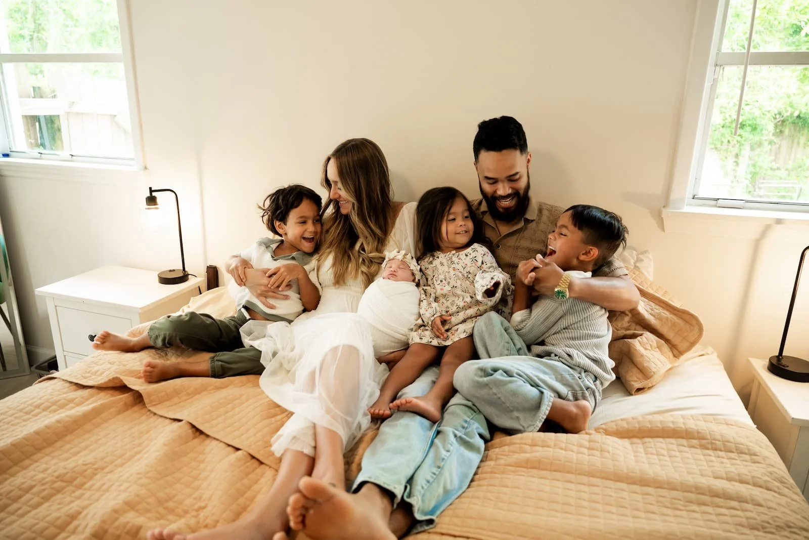 Family of six sitting on a bed in a bright bedroom, smiling and laughing together.