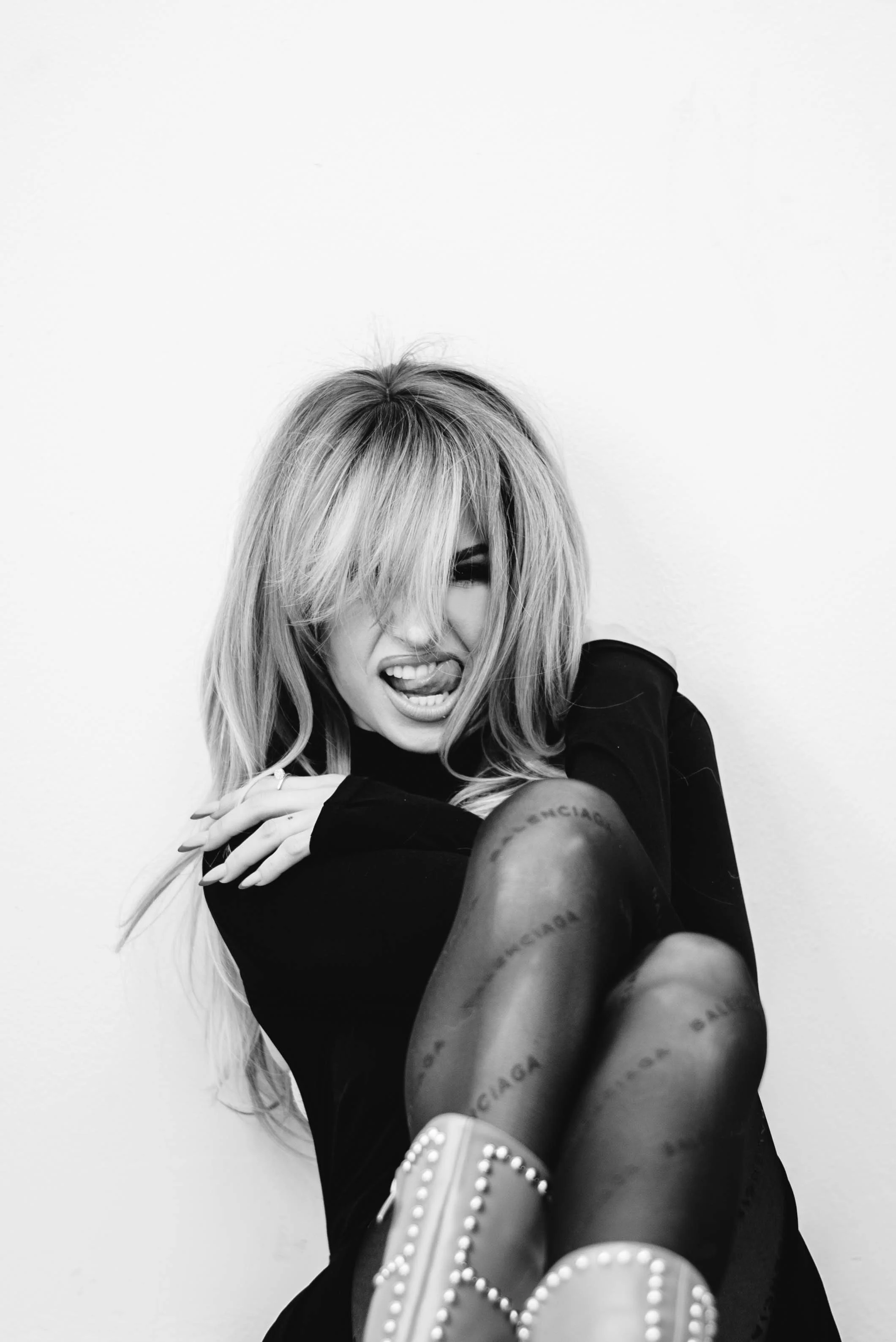 Black and white photo of a woman with tousled hair, showing her tongue and partially covering one eye, sitting against a plain wall, wearing a dark outfit and pattern tights with the word 'Balenciaga' on them, along with studded shoes.
