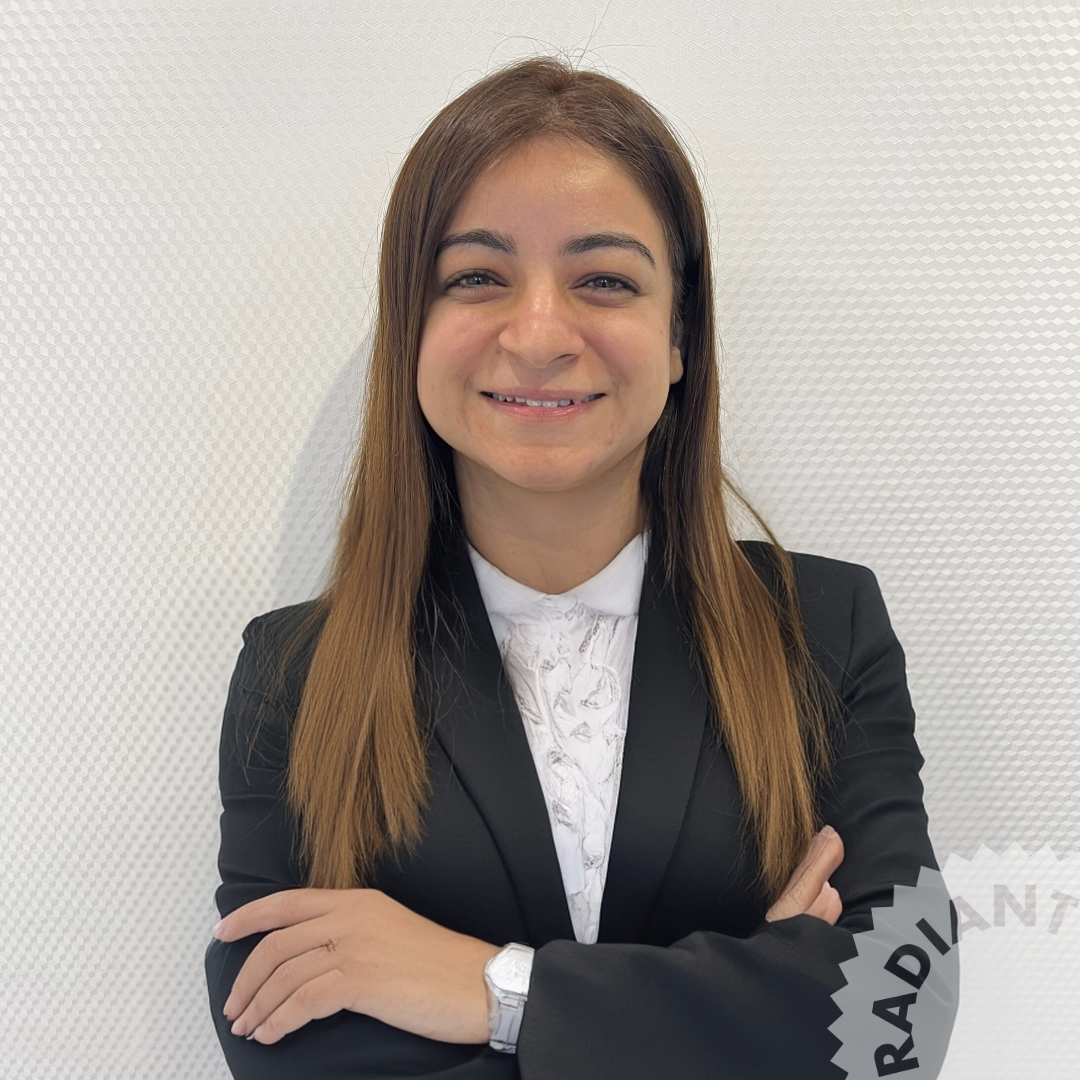 A woman with long brown hair in a professional black blazer and white blouse smiling with arms crossed, standing against a textured white wall.