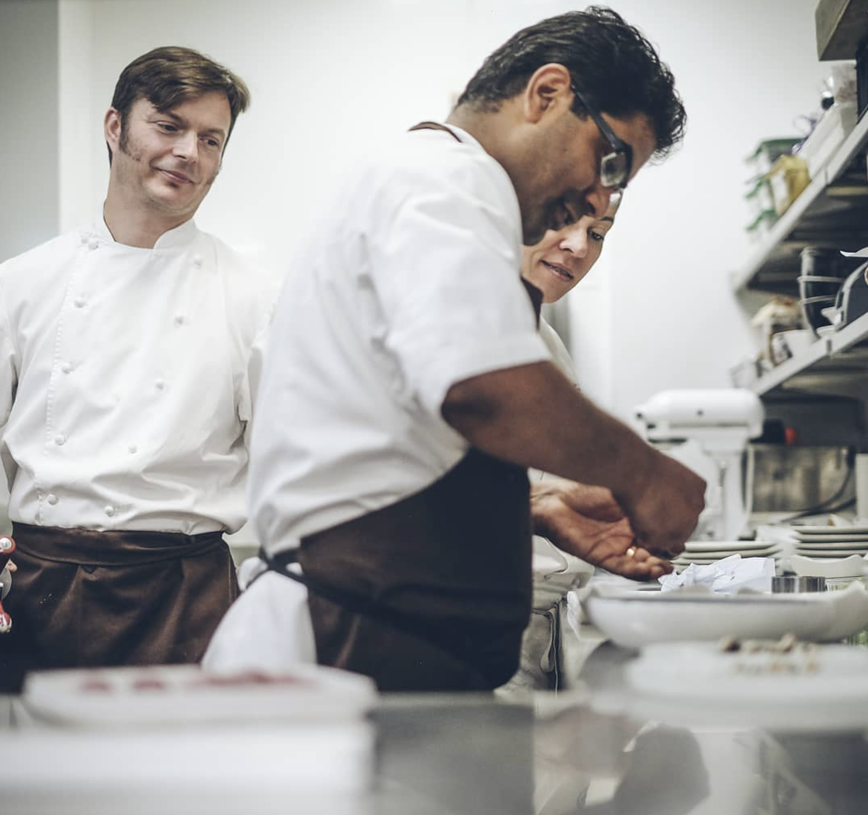 Three chefs in a commercial kitchen, with one actively preparing food and two observing. All are dressed in white chef jackets, with one wearing glasses and an apron.
