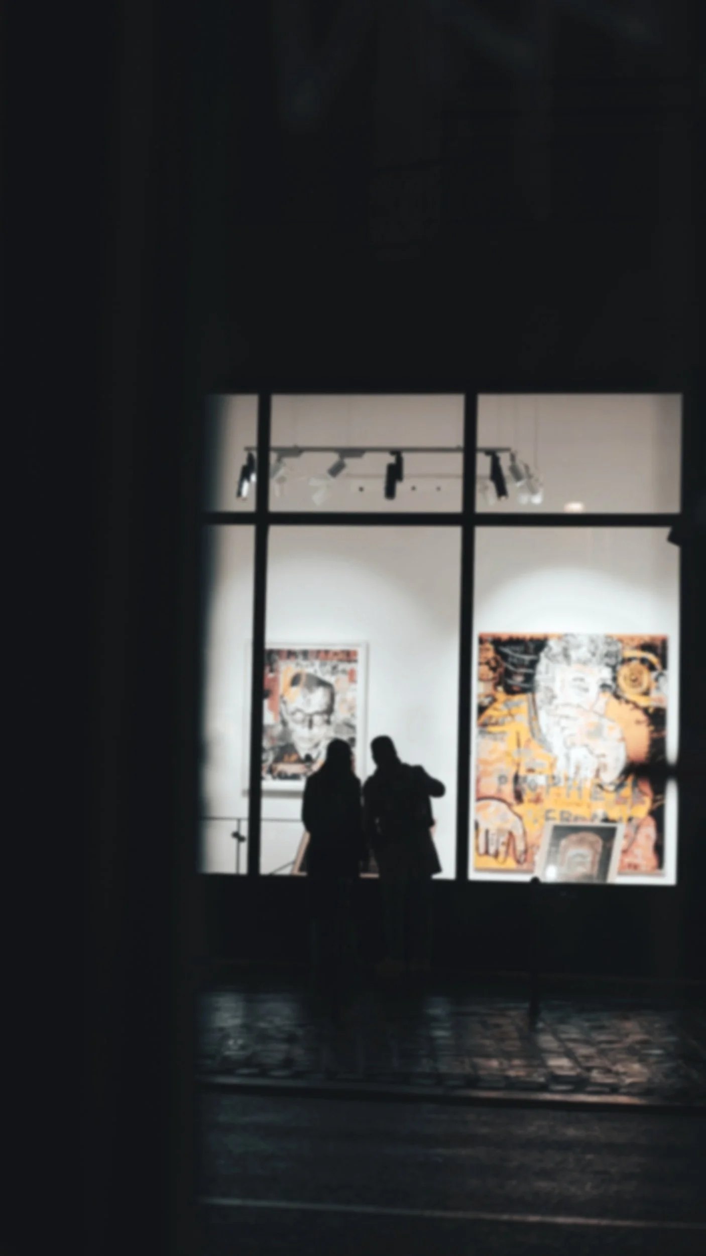 Silhouetted figures viewing artwork in an art gallery through a window.
