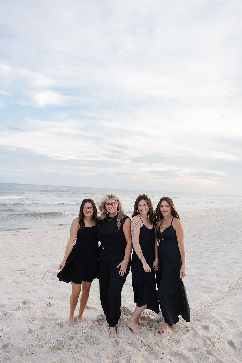 Four women standing barefoot on the sandy beach, smiling at the camera with the ocean and cloudy sky in the background.