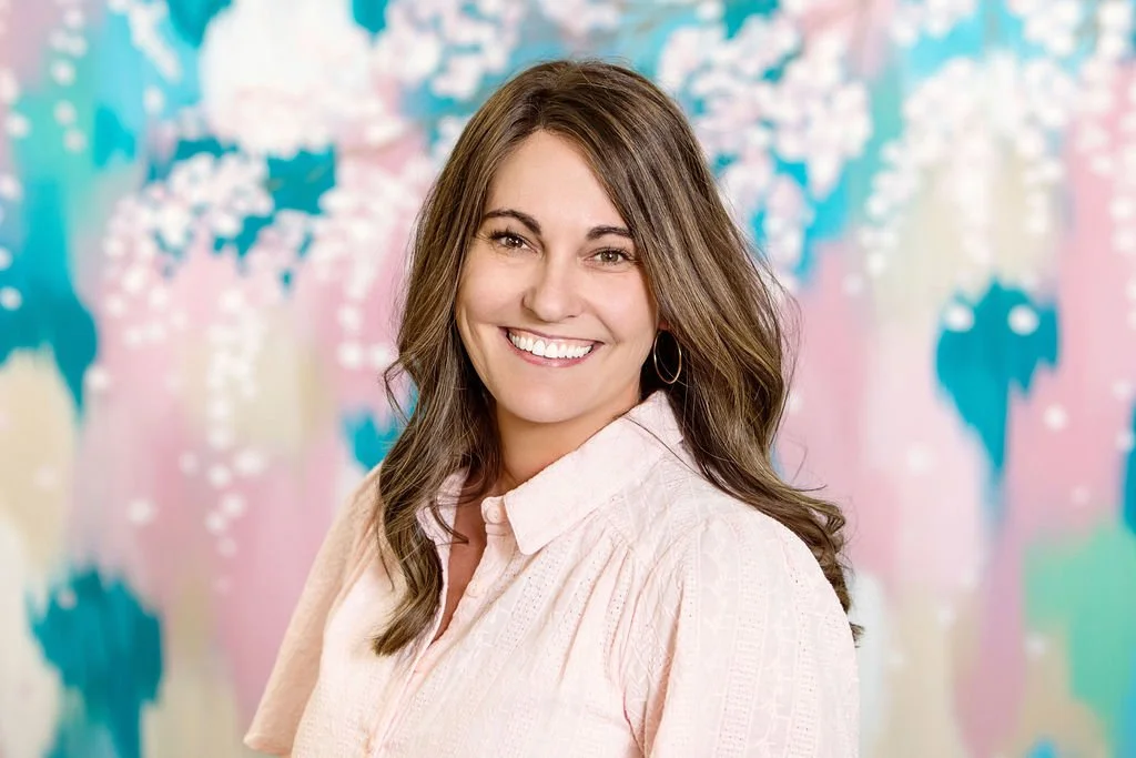 Portrait of a smiling woman with long brown hair wearing a light pink blouse, standing in front of a colorful abstract background.