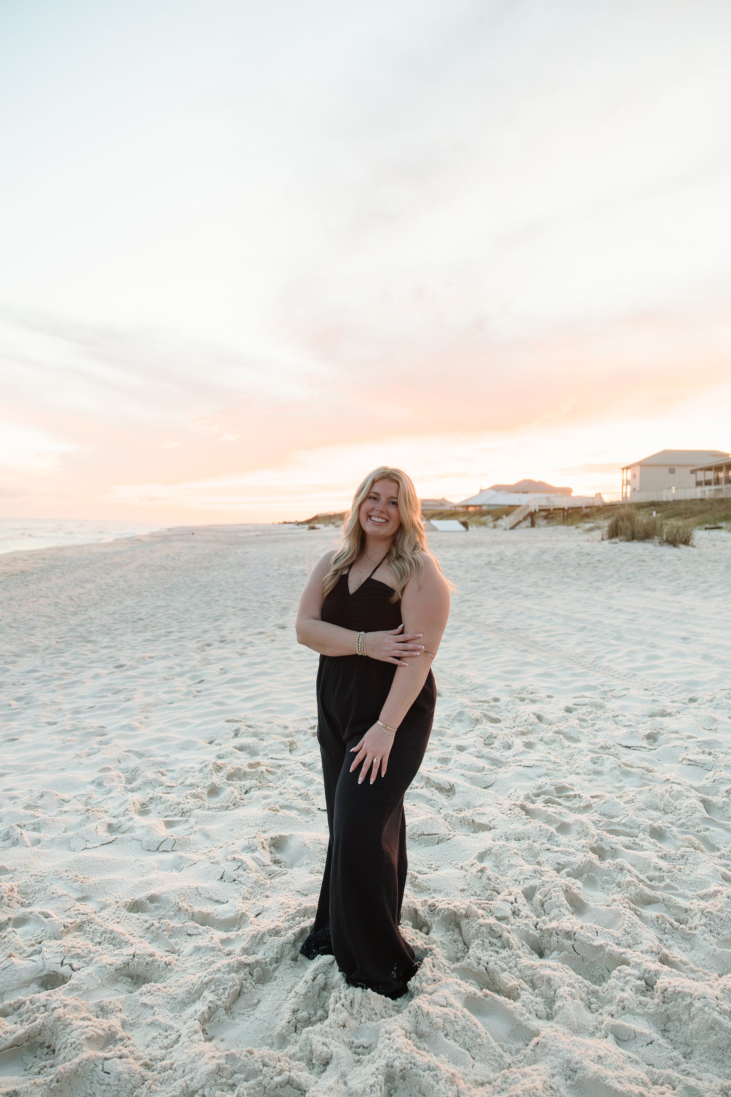 A smiling woman in a black dress standing on a sandy beach at sunset, with houses in the background and the sky painted with soft pastel colors.