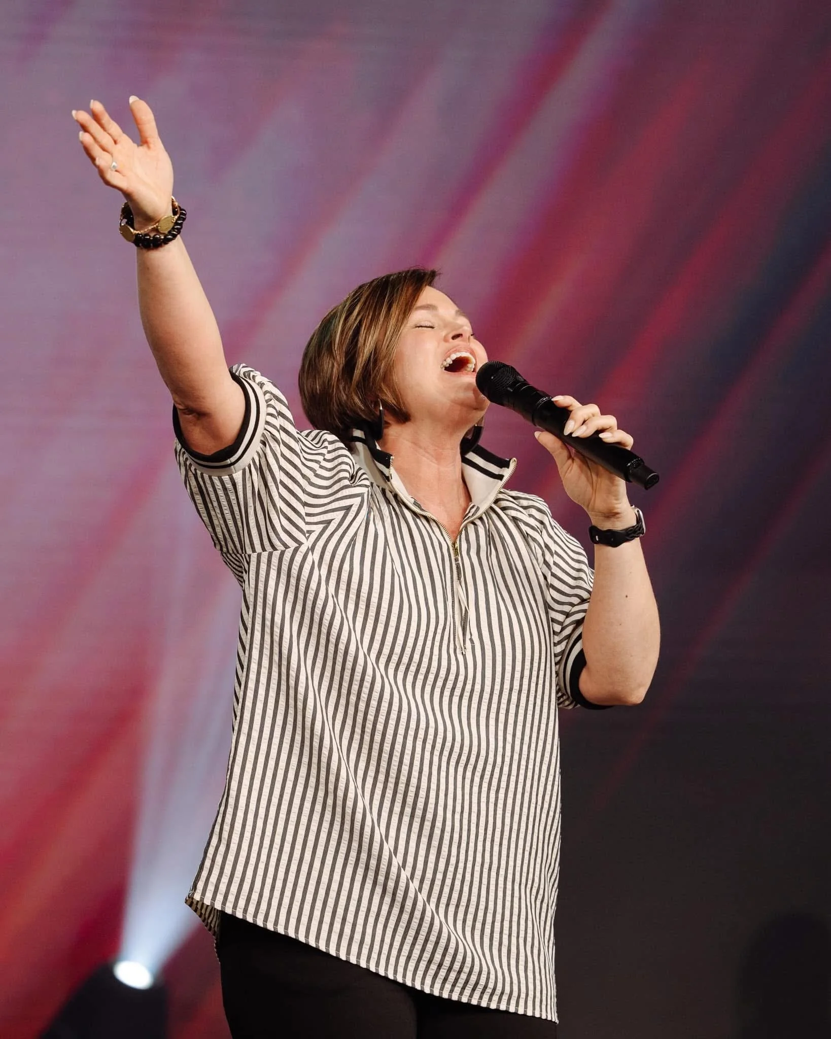 A woman with short brown hair singing passionately into a microphone with her eyes closed, wearing a striped shirt and bracelets on her wrist, against a blurred purple and red background.