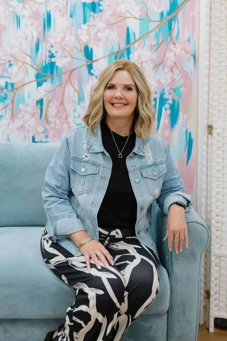 A woman with blonde hair wearing a cream sweater with textured pom-poms, blue jeans, layered bracelets, and a long necklace, standing in front of a colorful, abstract pink, blue, and brown painted backdrop with cherry blossom branch designs.