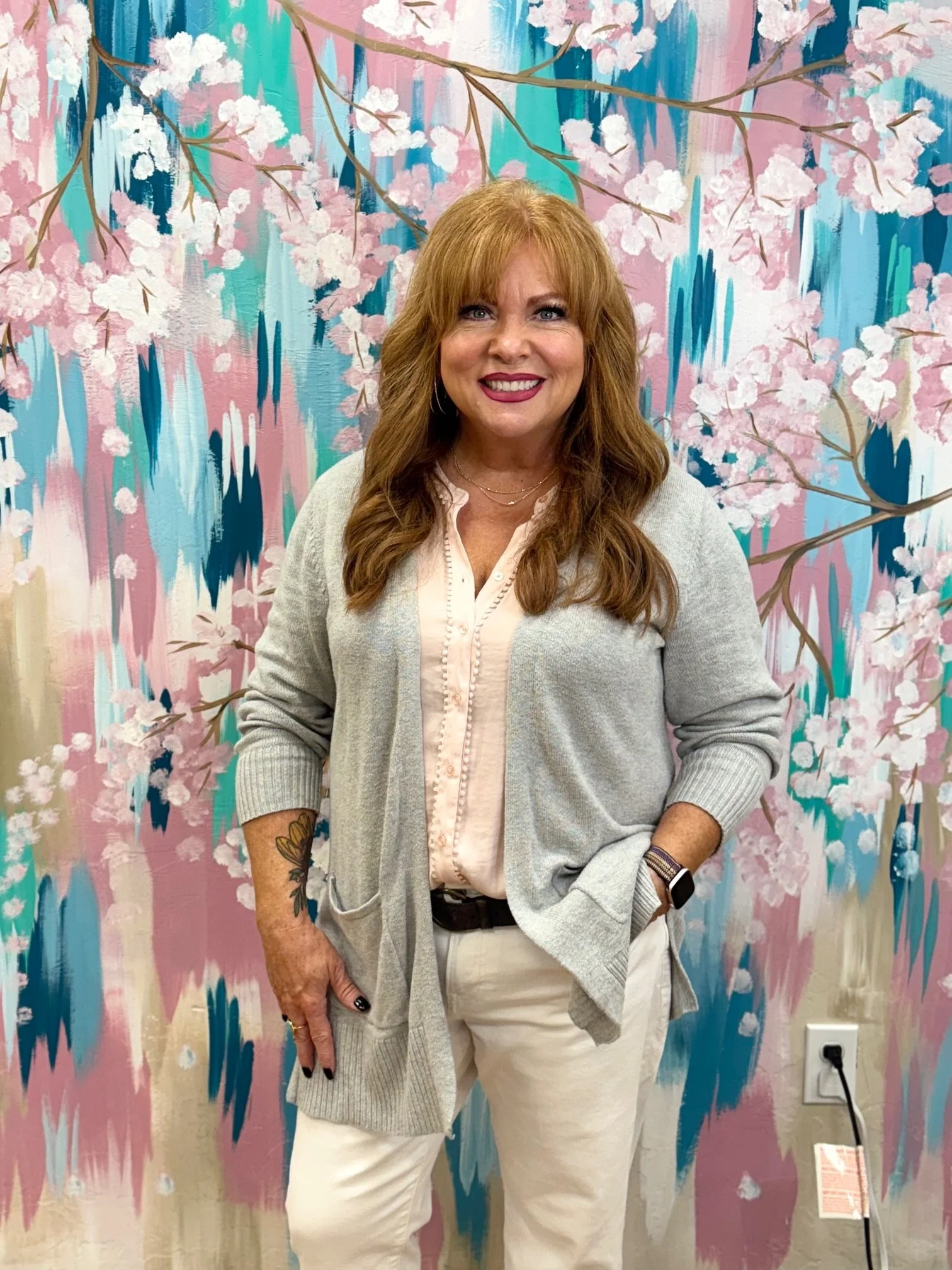 A woman with red hair smiling in front of a colorful, floral, painted wall with pink blossoms and blue, pink, and teal brush strokes.
