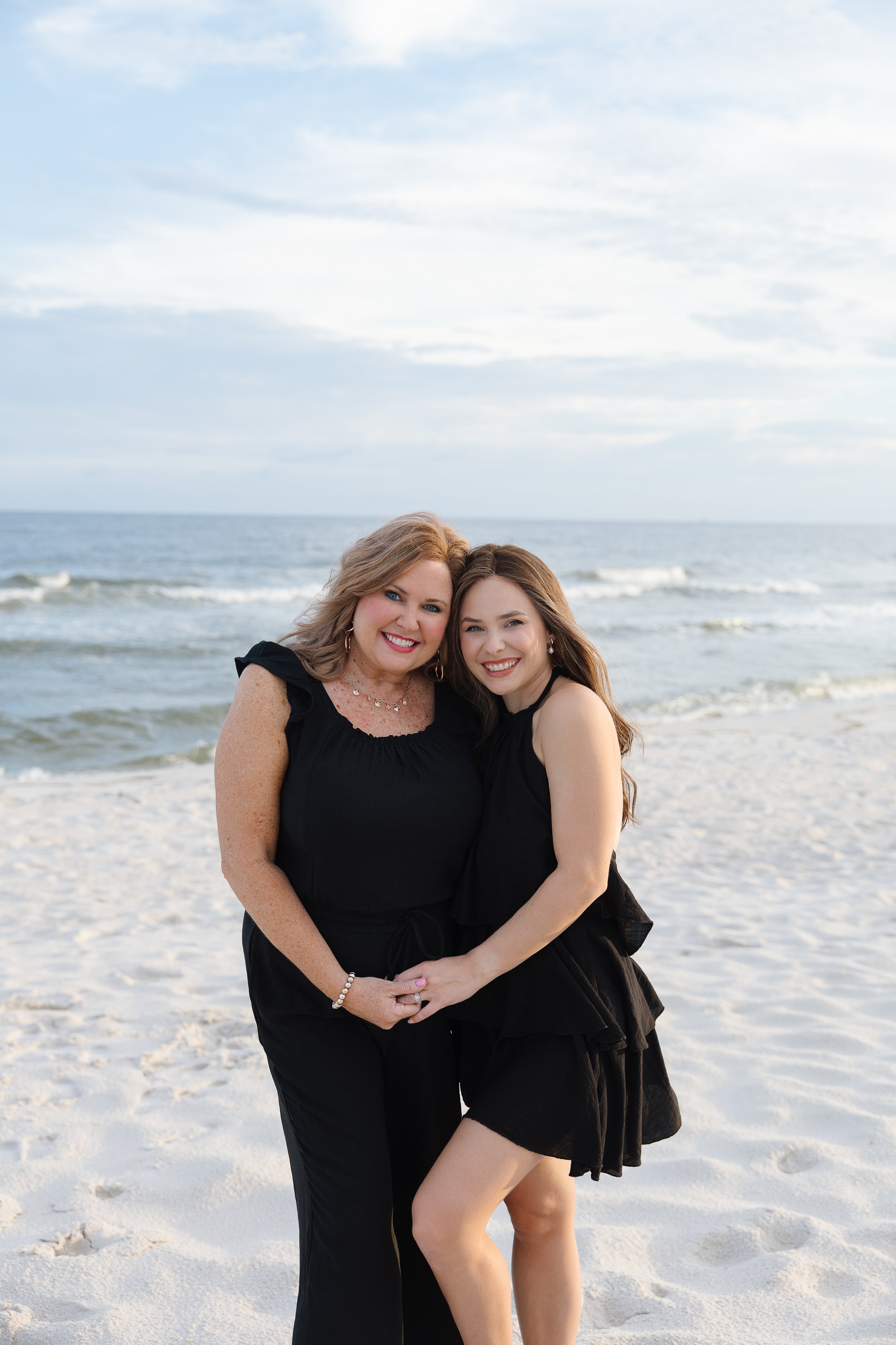 Two women standing close together on a beach with the ocean behind them, smiling at the camera.