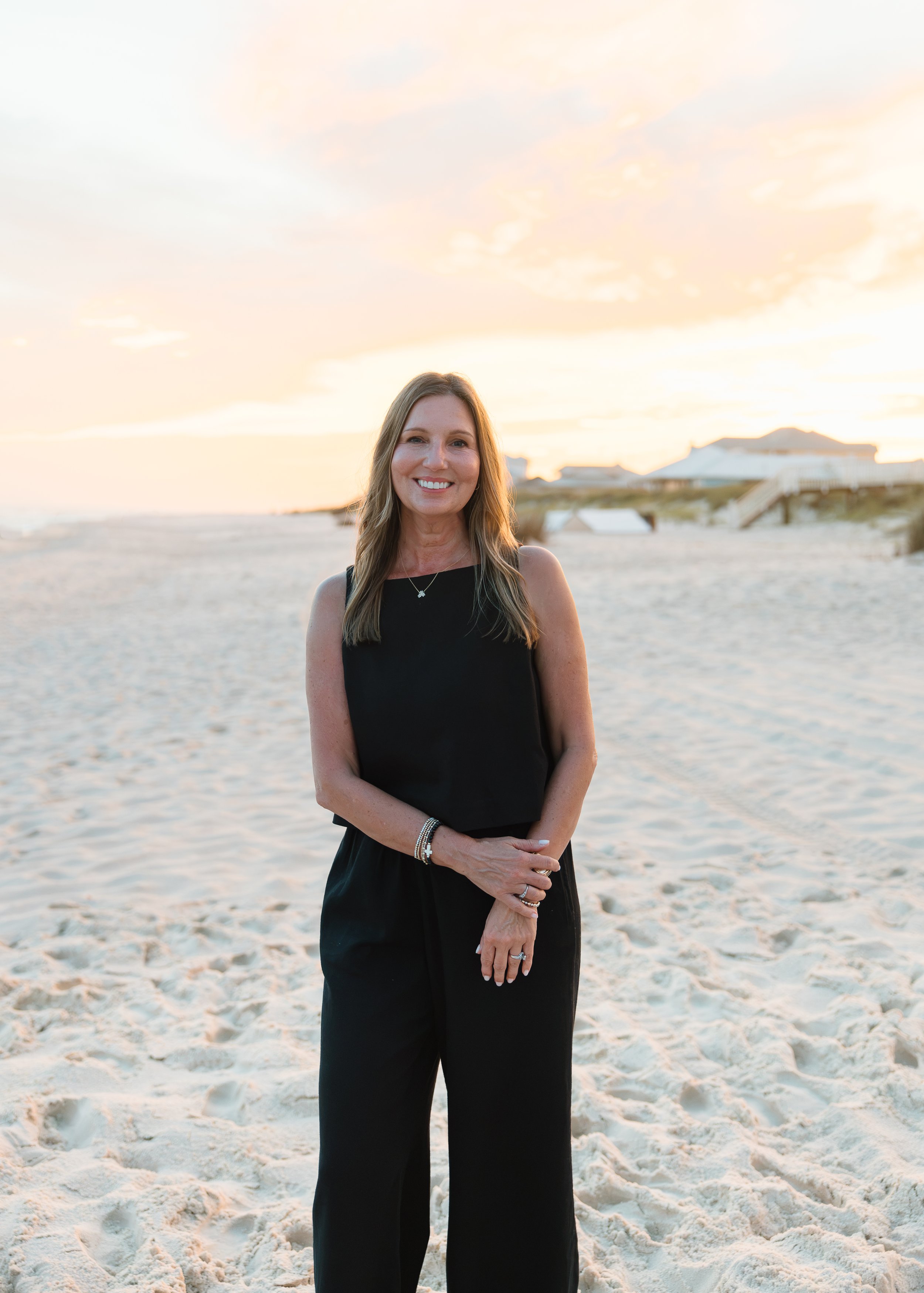 A woman with long light brown hair, smiling, wearing a black sleeveless top and black pants, standing on a sandy beach at sunset with houses and boardwalks in the background.