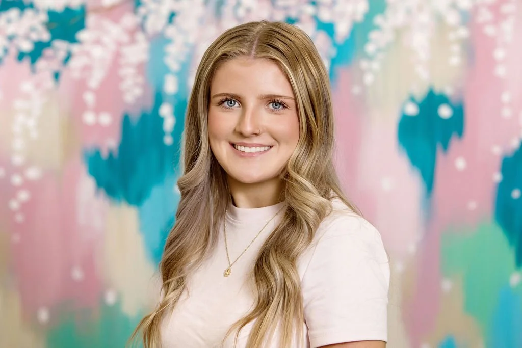 A young woman with long, wavy blonde hair smiling at the camera against a colorful, abstract background with blue, pink, and purple shapes.