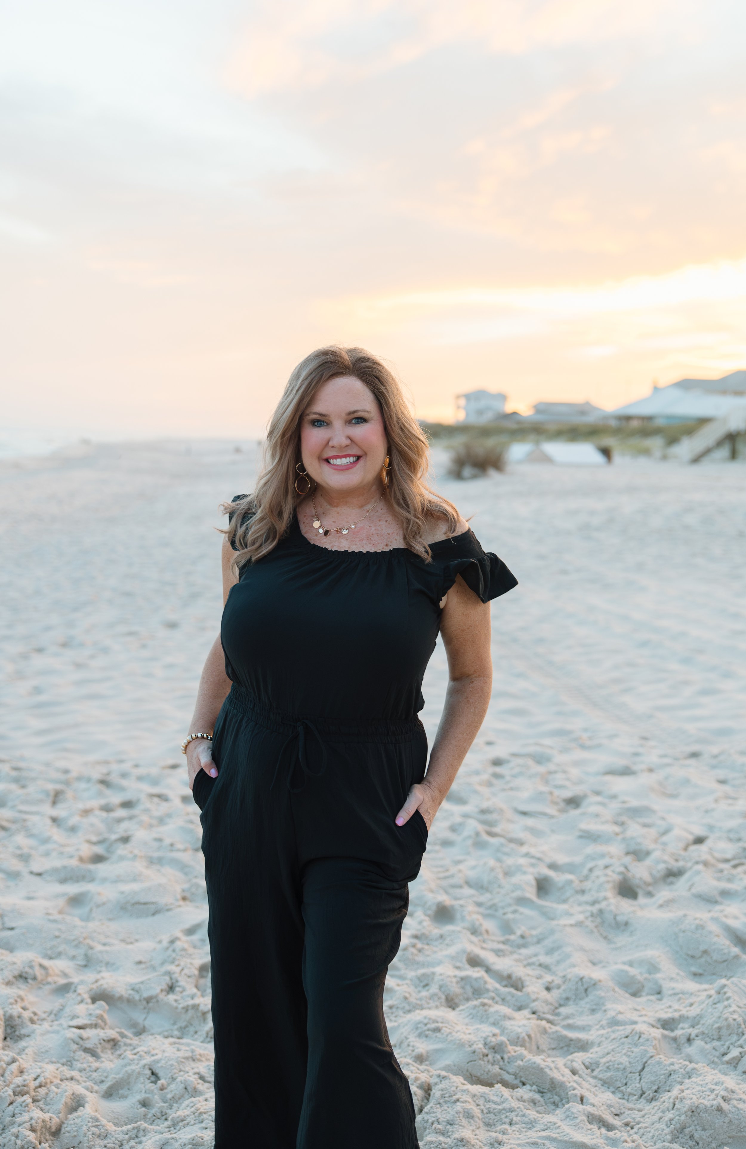 A woman with blonde hair and jewelry, wearing a black dress, standing on a sandy beach at sunset, smiling at the camera.